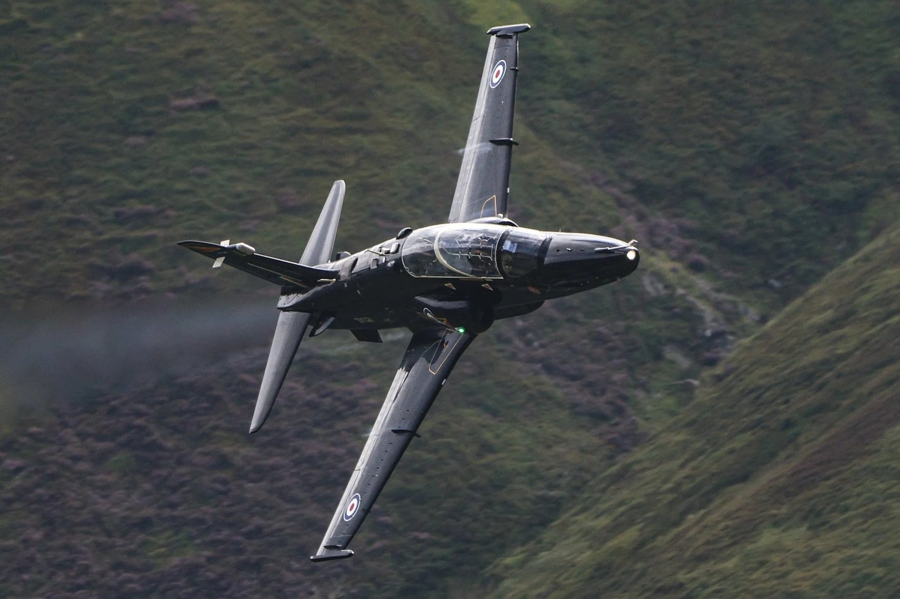 A jet airplane flying over a green, mountainous landscape.