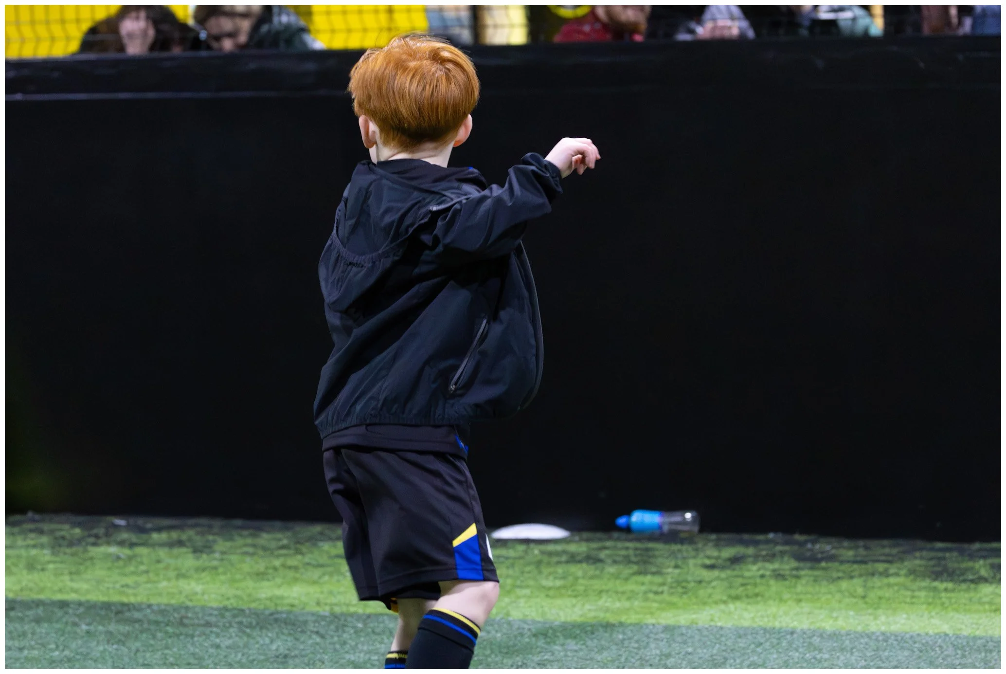 A young boy with red hair wearing a black jacket and shorts is standing on a green surface, possibly a sports field, with a black barrier in the background. There are spectators visible behind the barrier.