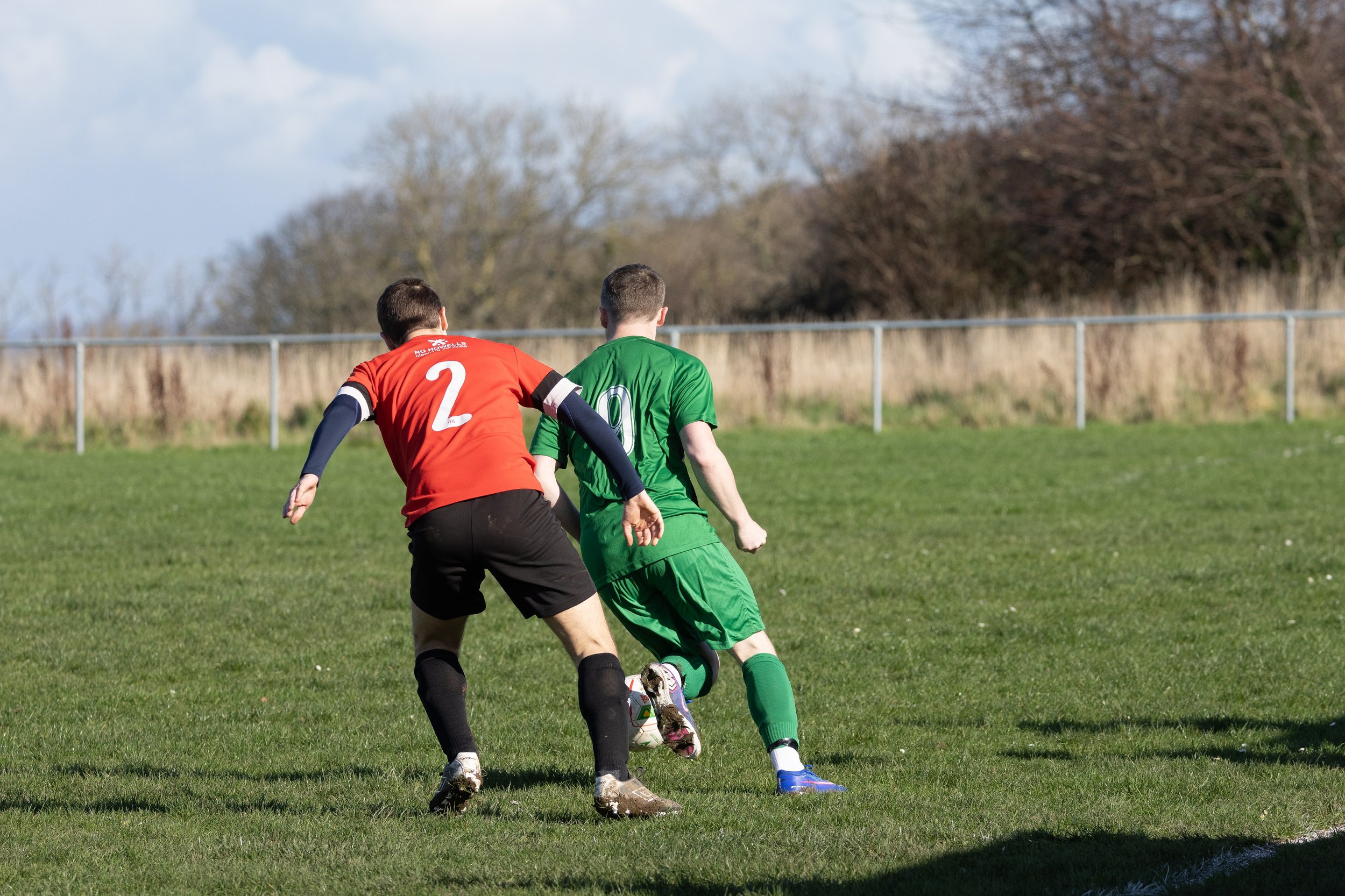 Two soccer players, one in a red jersey and the other in a green jersey, playing on a grassy field during daytime.