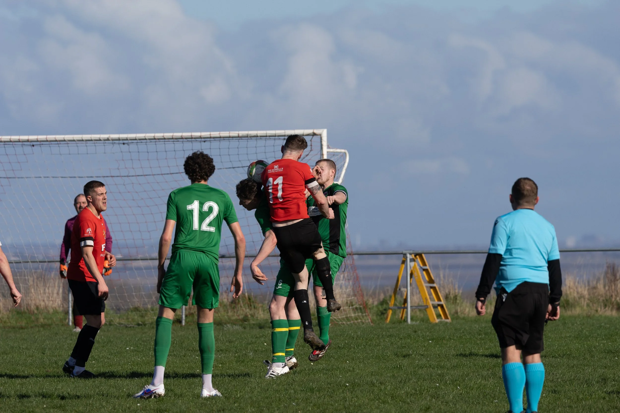Soccer players in green and red uniforms jumping and positioning near the goal on a grassy field during a game, with a referee watching.