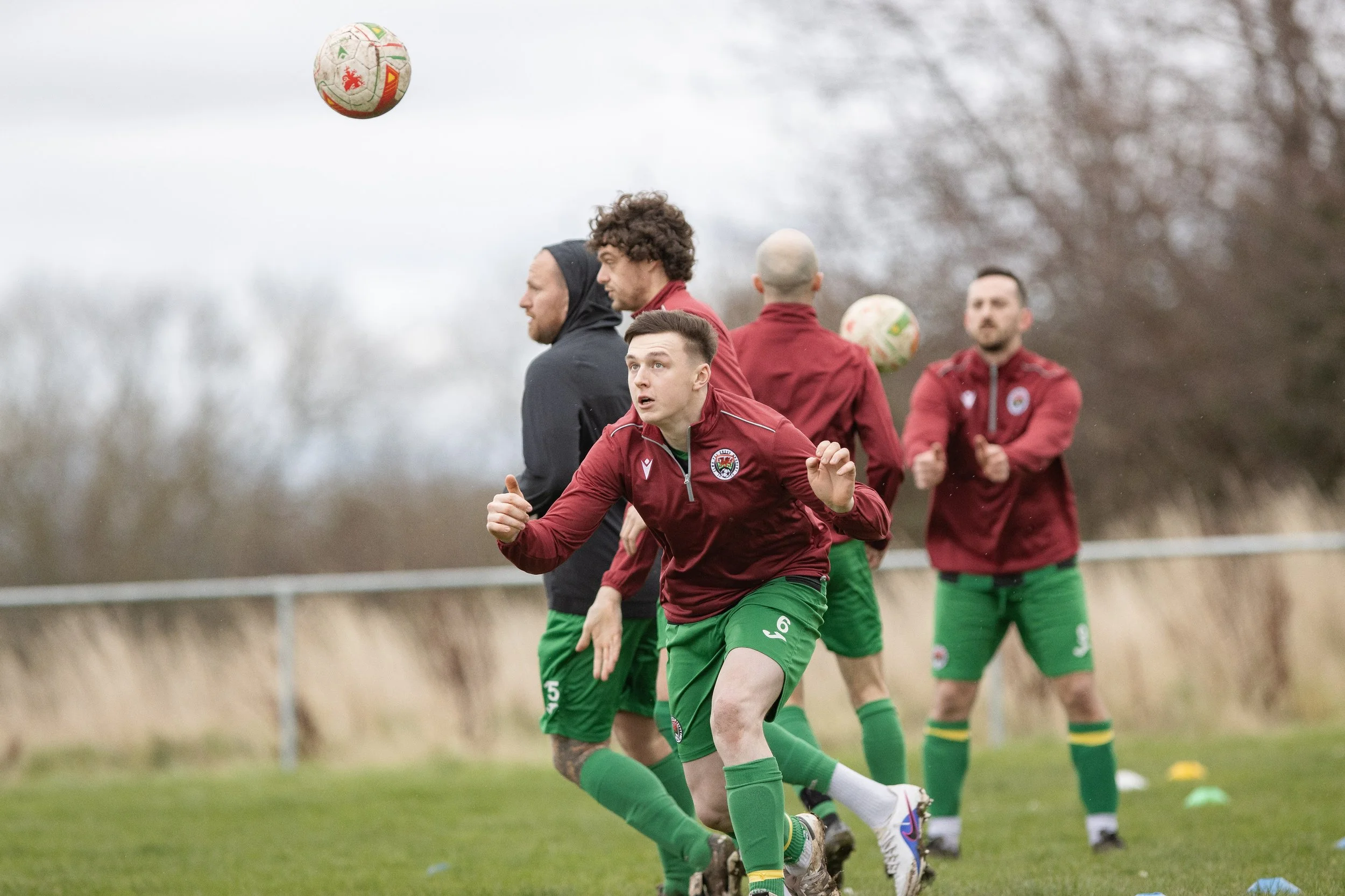 Soccer players in red and green uniforms practicing on the field, with two soccer balls in the air and a background of trees.