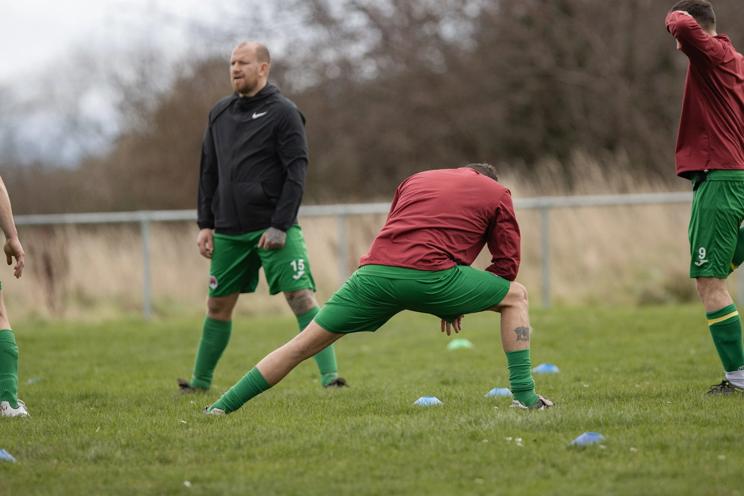 Soccer players on a field during training or practice, one stretching with a wide stance and bent over, others standing nearby, with a coach or observer watching in the background.