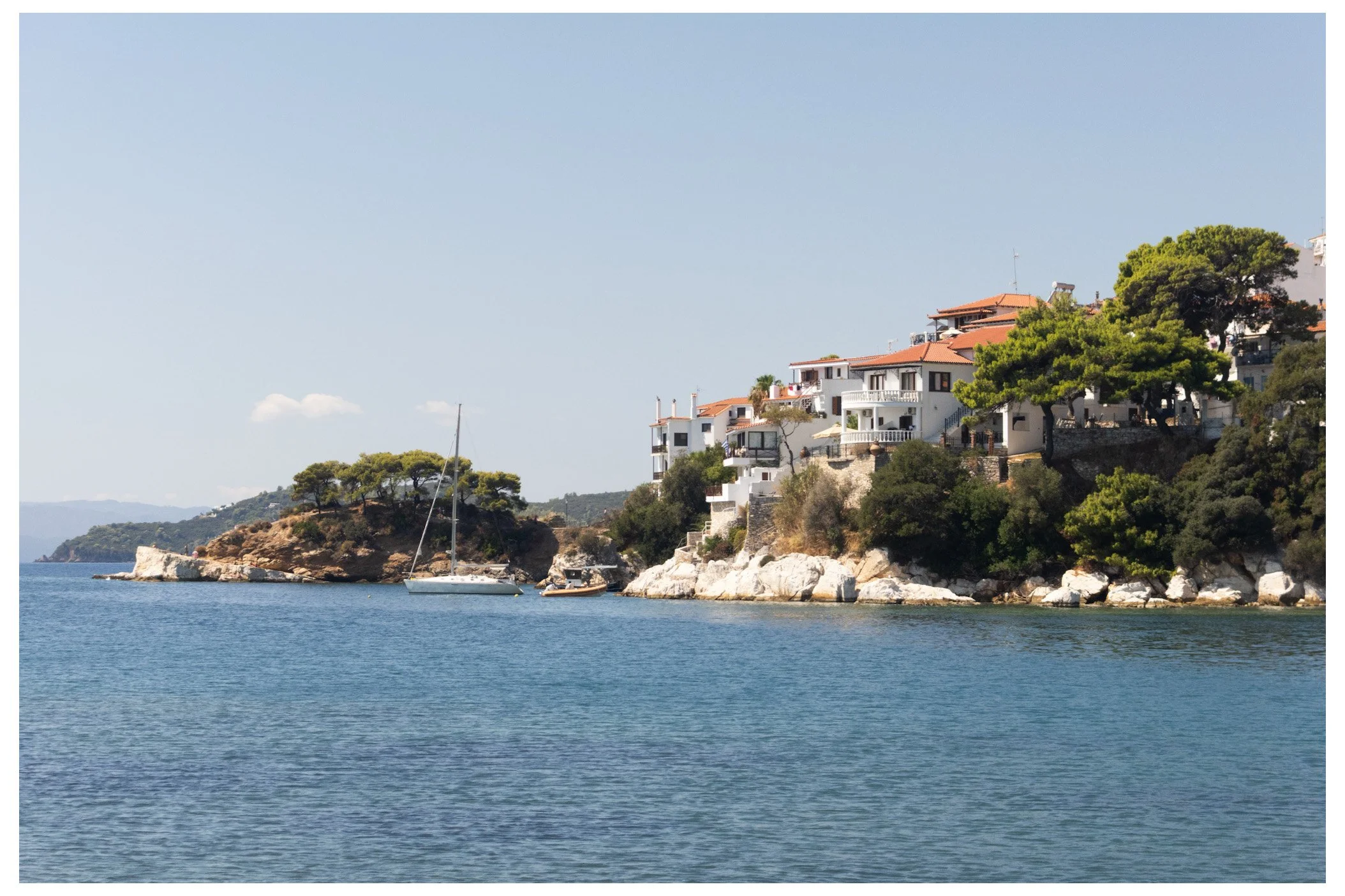 A coastal scene with houses on a hillside, trees, rocks, and a sailboat in the water.