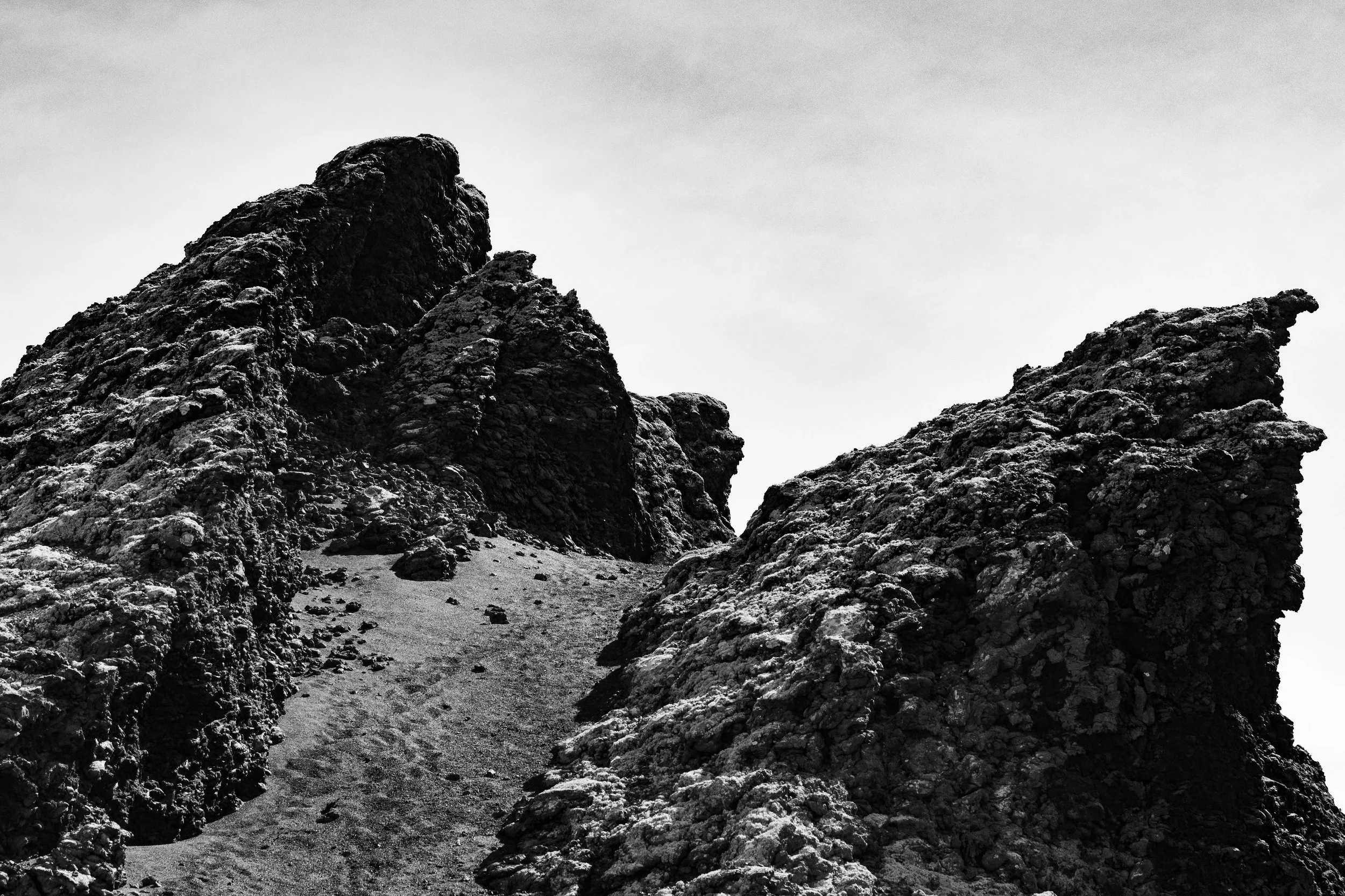 Black and white photo of large, rugged volcanic rocks with a sandy pathway between them, under a cloudy sky.