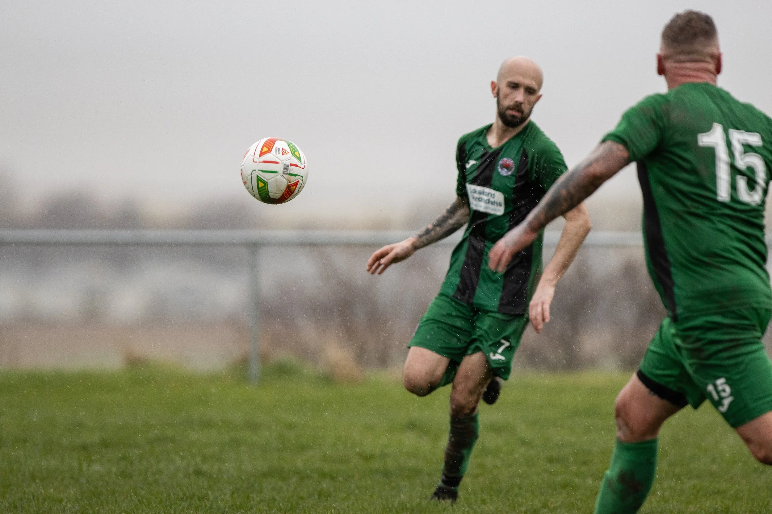 Two soccer players wearing green jerseys playing in the rain; one is kicking a white soccer ball with red and green accents.