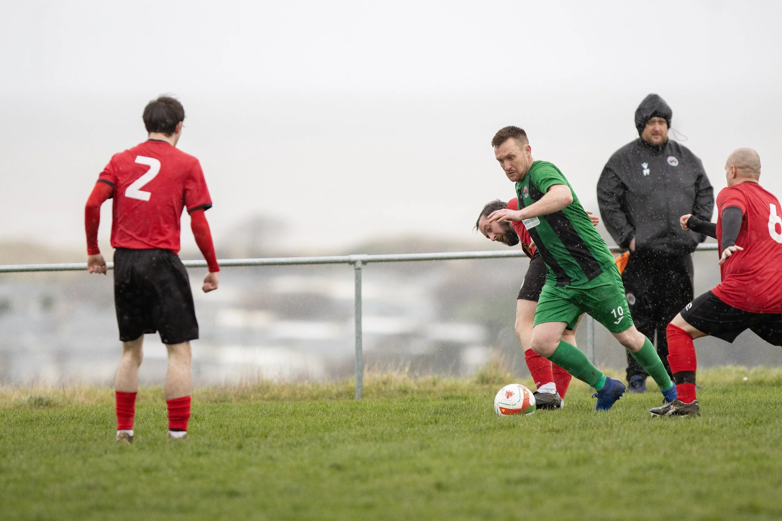 Soccer players in red and green jerseys compete for the ball on a rainy day.