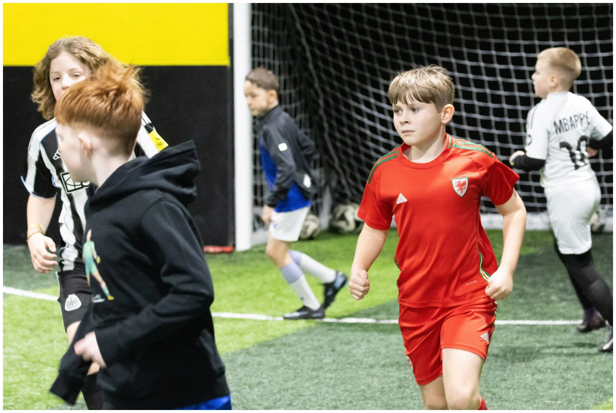 Group of young boys playing indoor soccer, with one boy in a red jersey in the foreground, others in black and white jerseys, and goal net in the background.