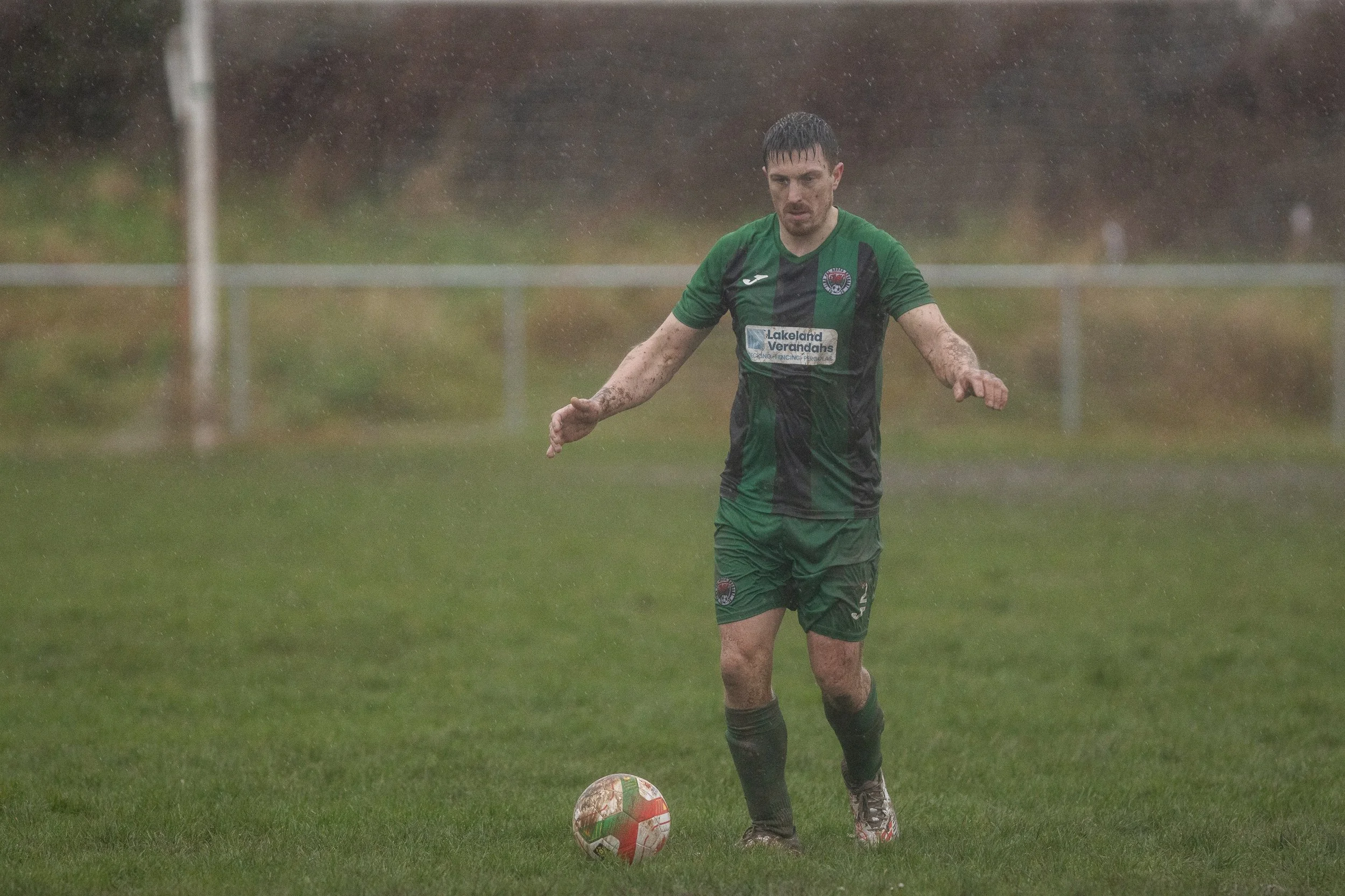 A soccer player wearing a green and black uniform is on a muddy field in the rain, about to kick a soccer ball.