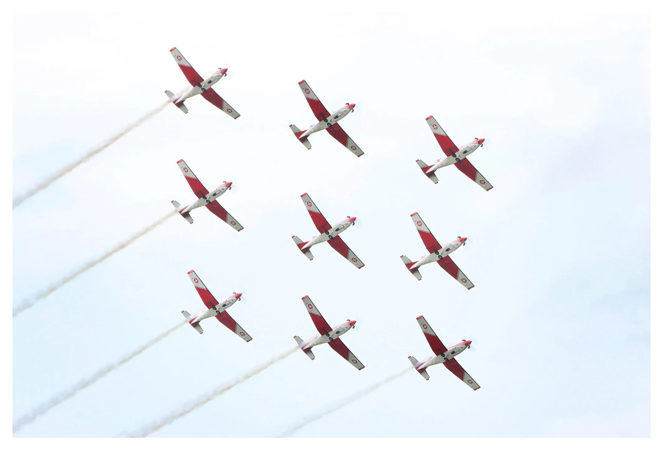 A formation of ten red and white planes flying in a tight diamond shape, leaving white smoke trails against a cloudy sky.
