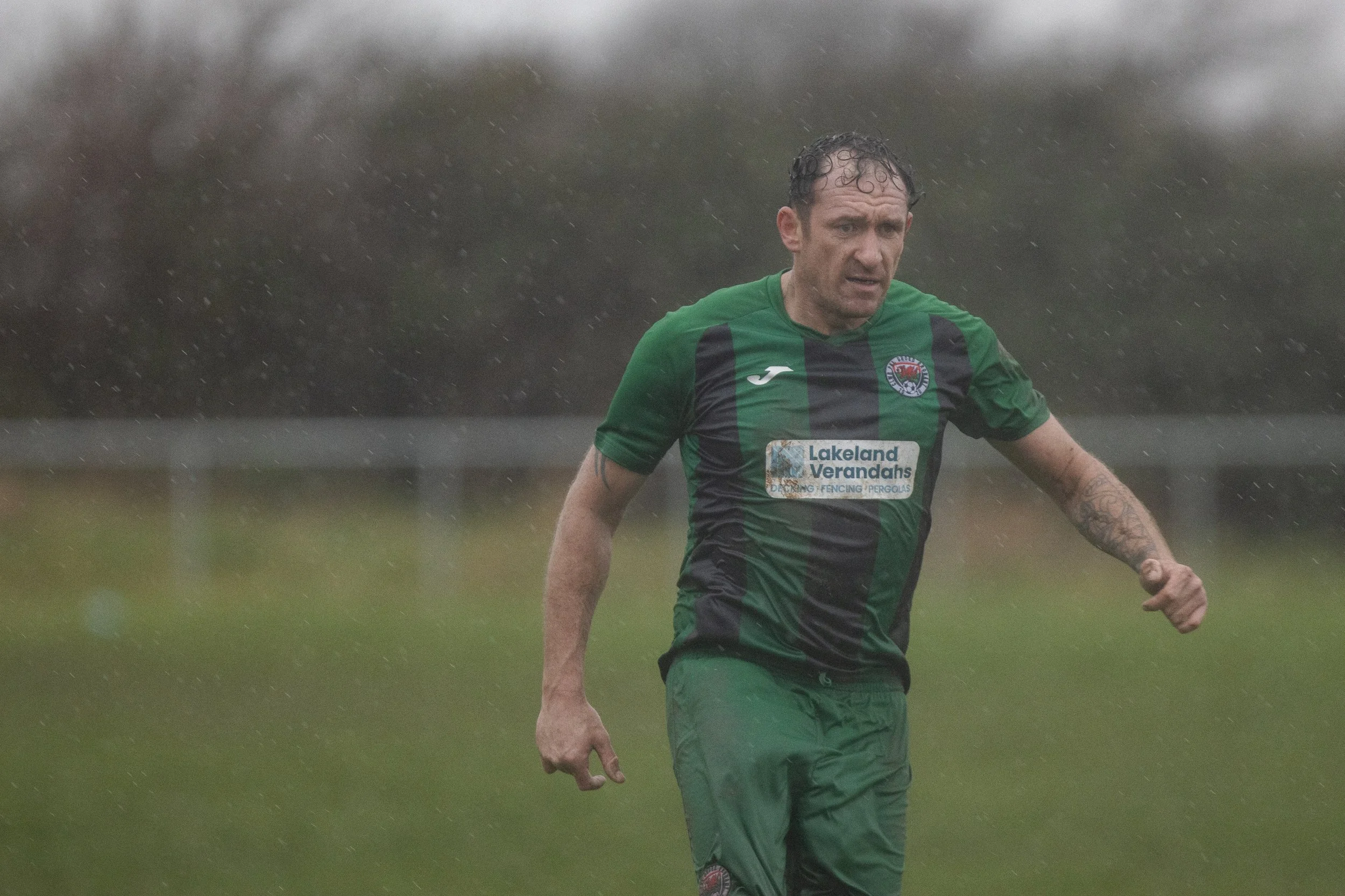 A man in a green and black soccer uniform running outdoors in the rain, with a blurred background of trees and grass.