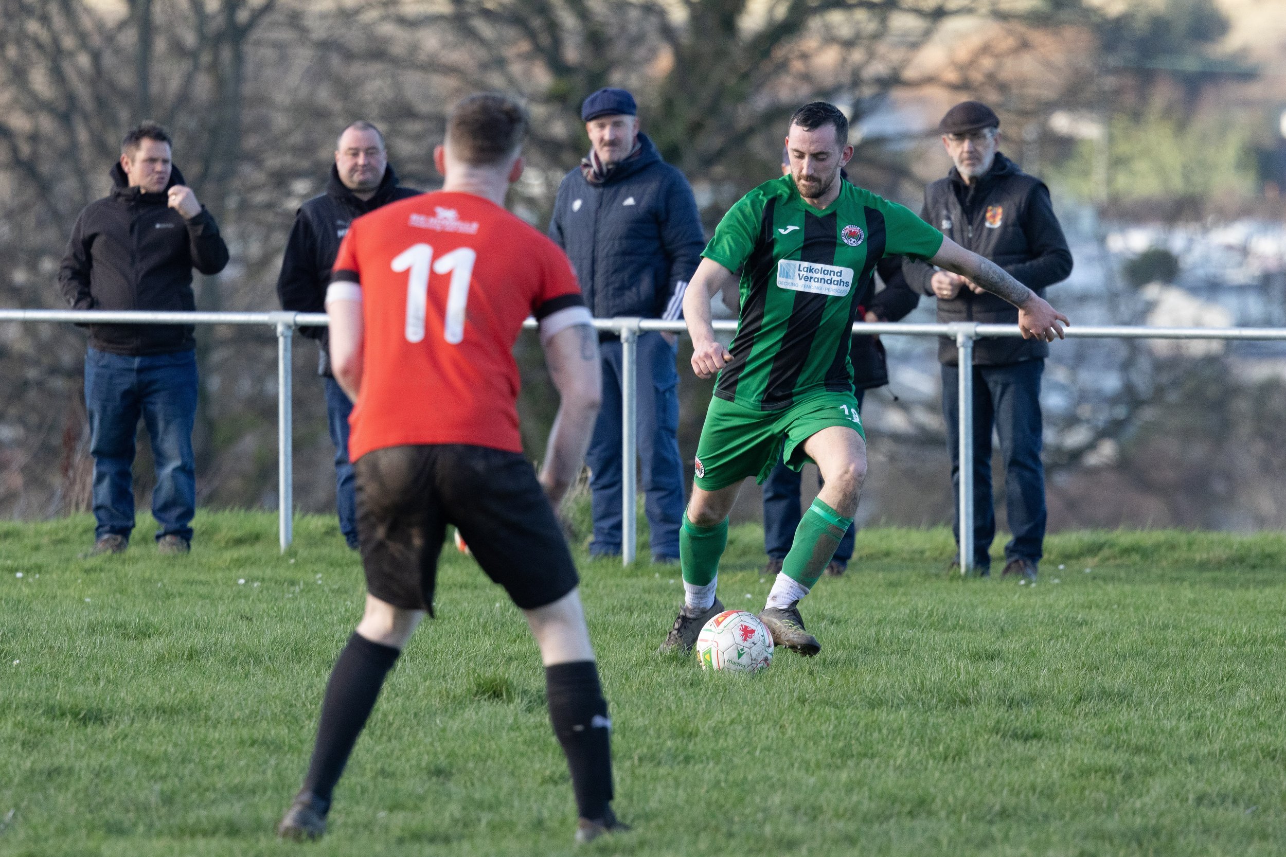 A soccer player in a green and black uniform preparing to kick a ball while another player in a red and black uniform faces him on a grassy field. Several spectators stand behind a metal barrier in the background.