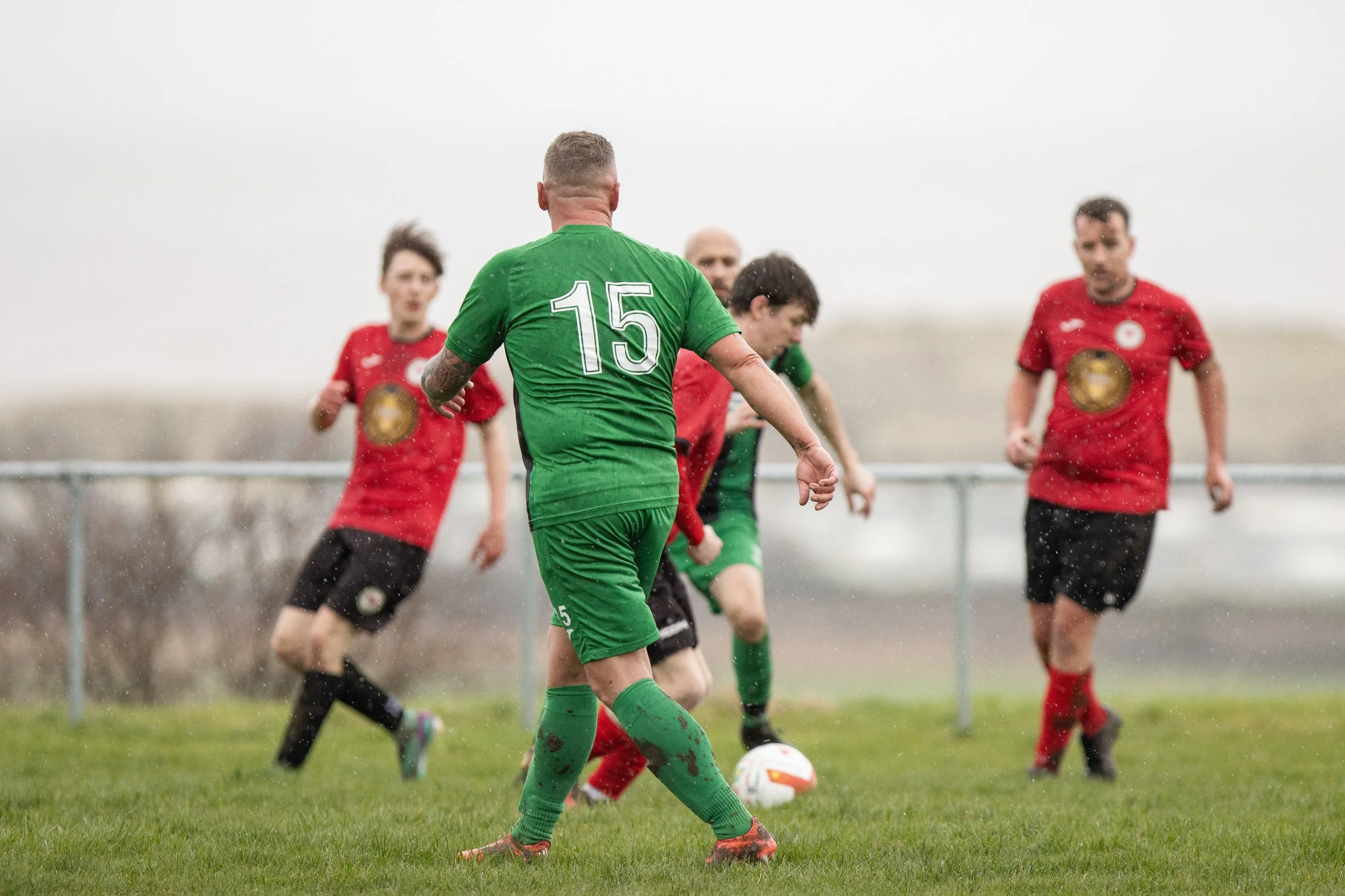 A soccer match in progress outdoors on a rainy day, with players in green and red jerseys competing for the ball.