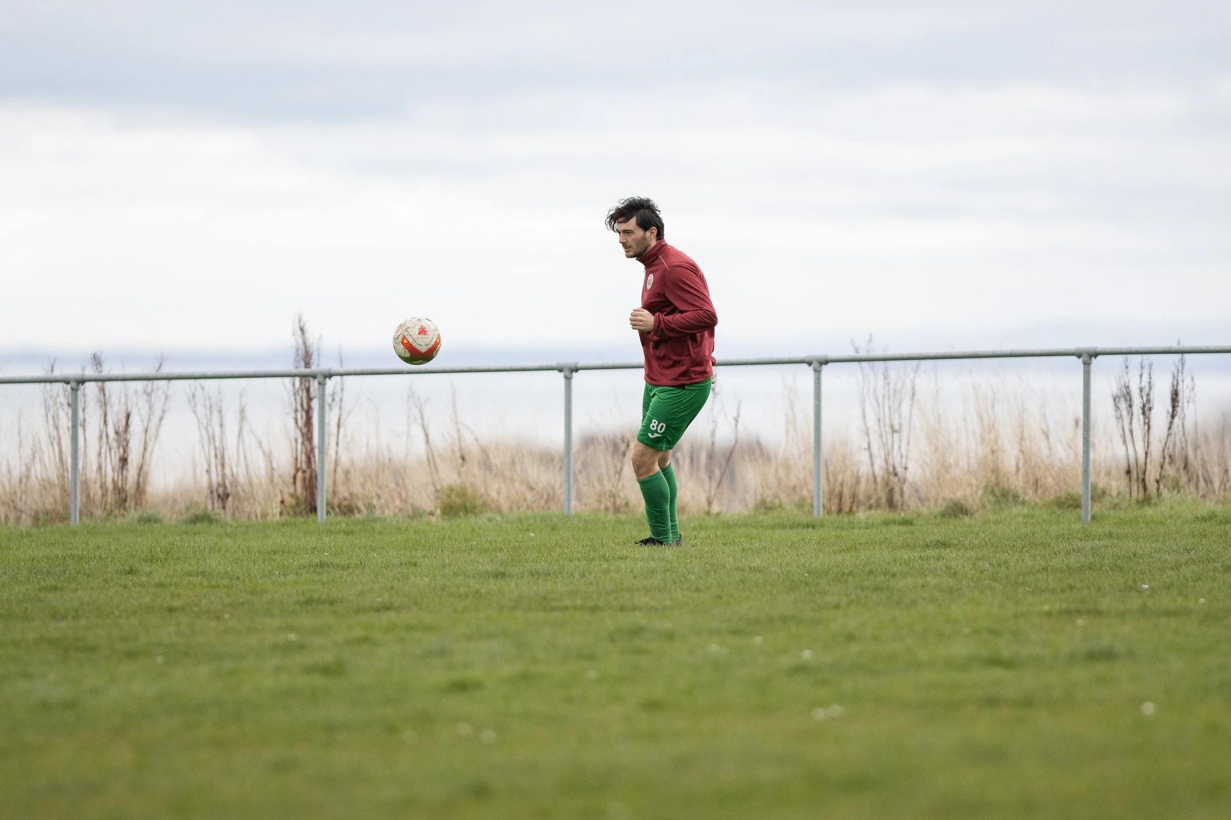 A man dressed in red and green soccer gear is on a grassy field, preparing to kick a soccer ball. The sky is overcast, and there is a metal railing and dried grass in the background.