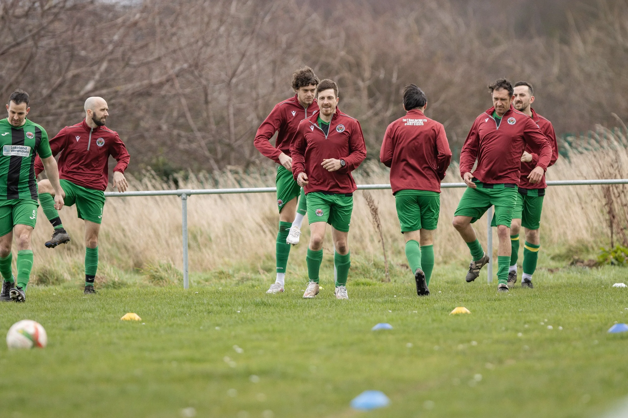 Group of male soccer players in training, wearing green shorts and maroon jackets on a grassy field with cones.