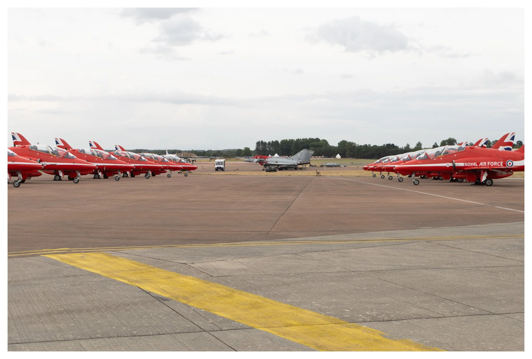 Multiple red training jets with Royal Air Force markings parked on an airfield tarmac, with a gray fighter jet and trees in the background.