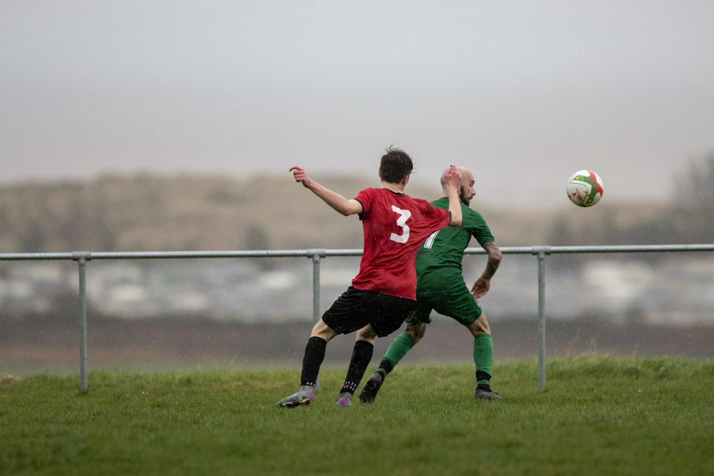 Two soccer players, one in a red jersey with number 3 and the other in a green jersey, competing for the ball during a game on a grassy field on a cloudy day.