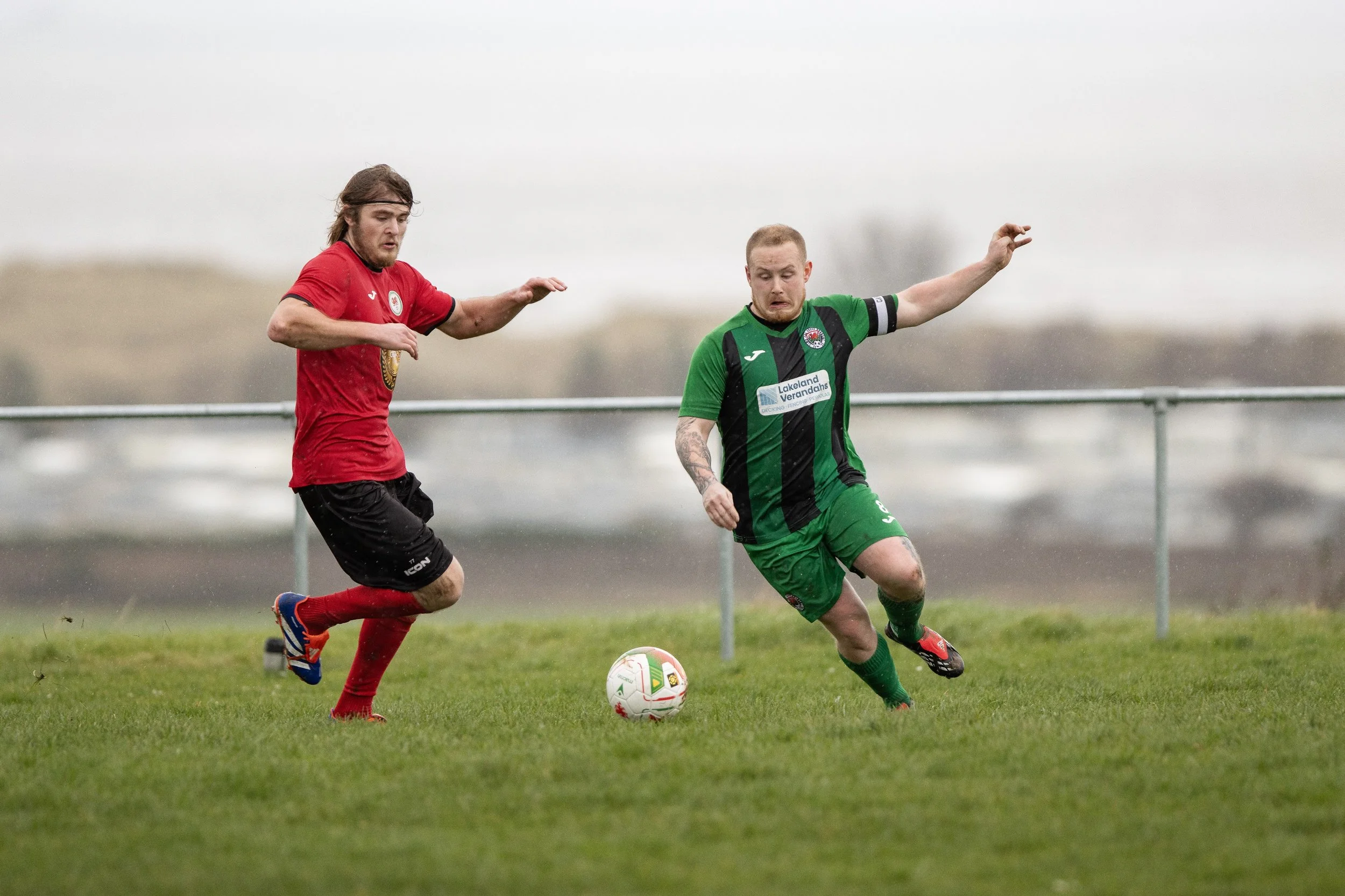 Two soccer players in red and green uniforms compete for the ball on a grassy field under gray skies.