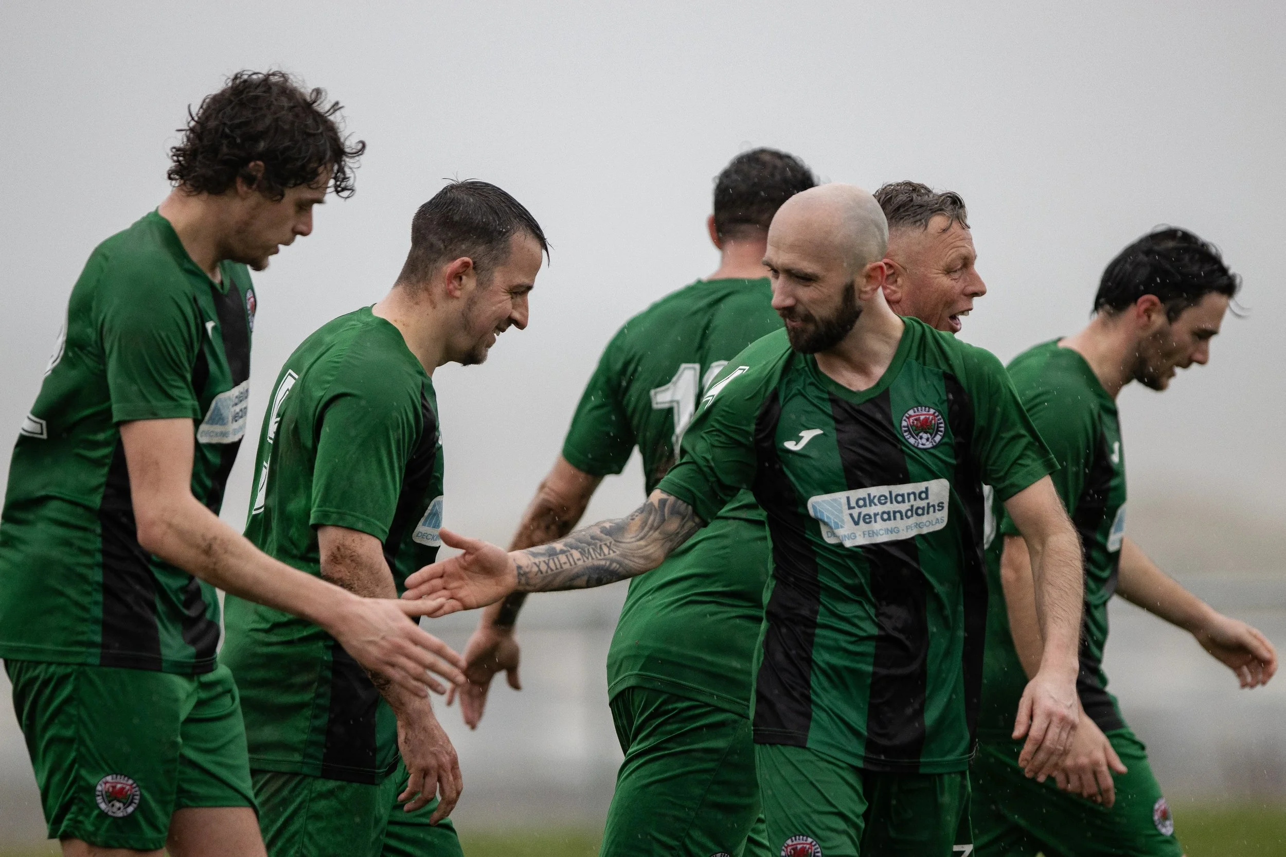 Soccer players in green jerseys high-five and celebrate on a field in rainy weather.