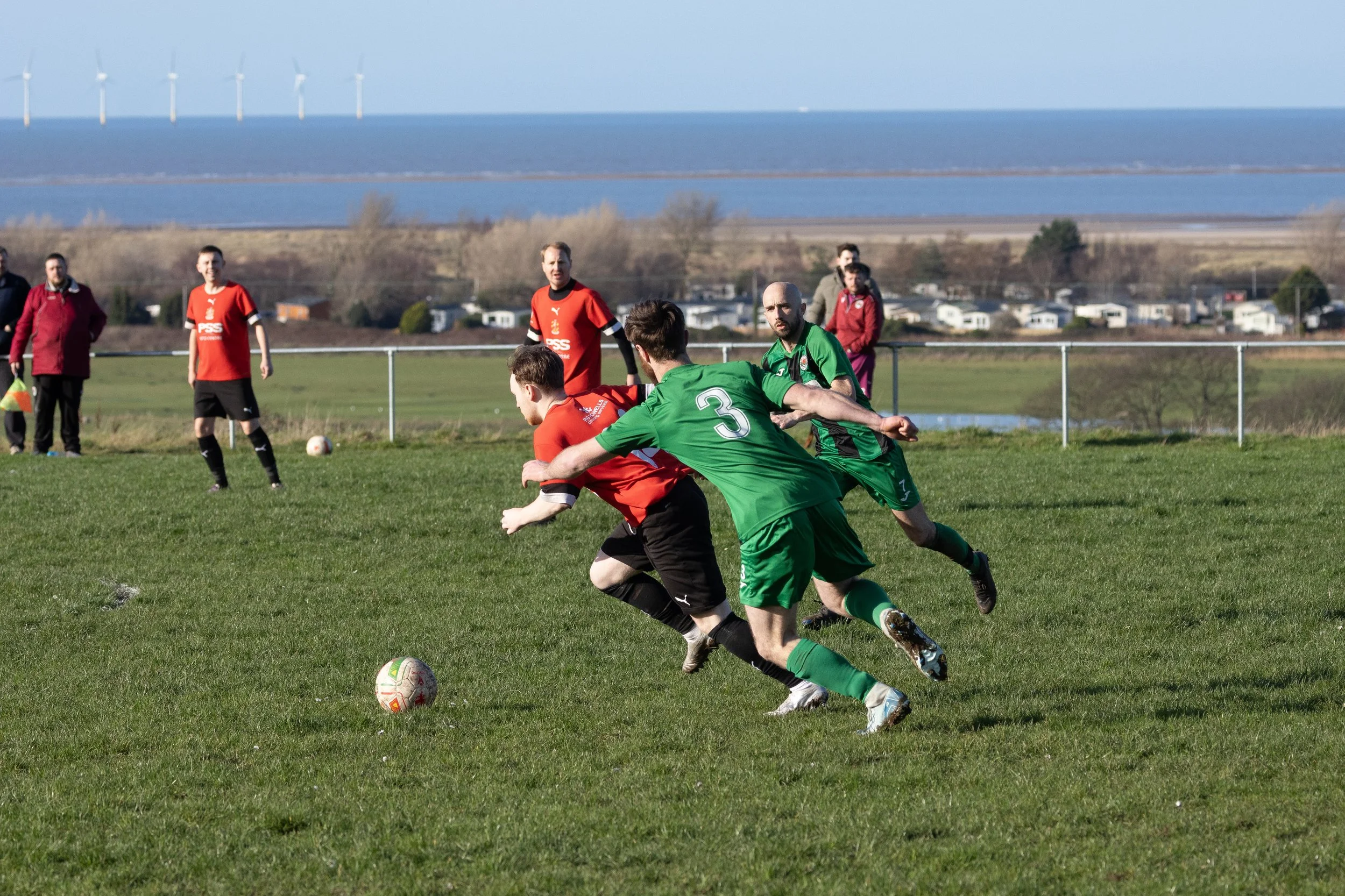 Soccer players in green and red uniforms competing for the ball on a grassy field with a scenic backdrop including water, wind turbines, and a small town.