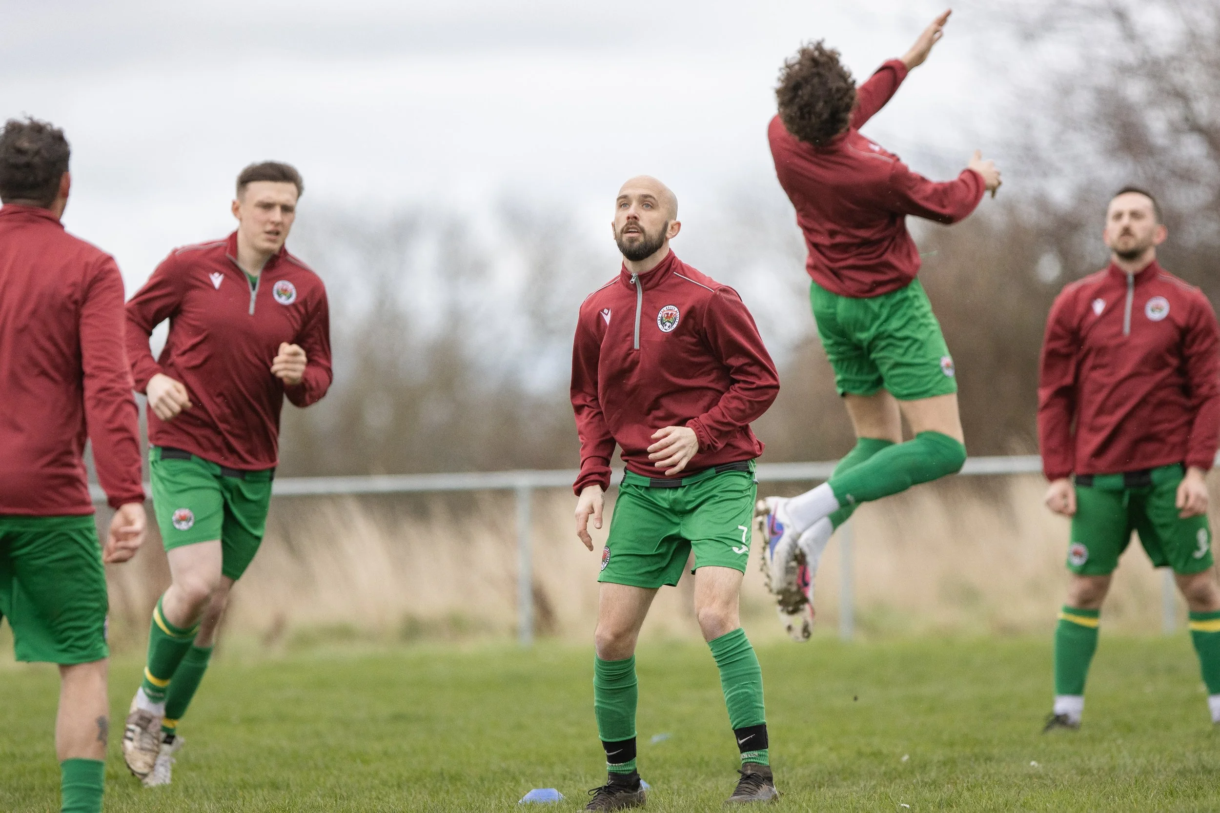Group of male soccer players in maroon jackets and green shorts warming up or practicing on a grassy field, some jogging and one jumping.