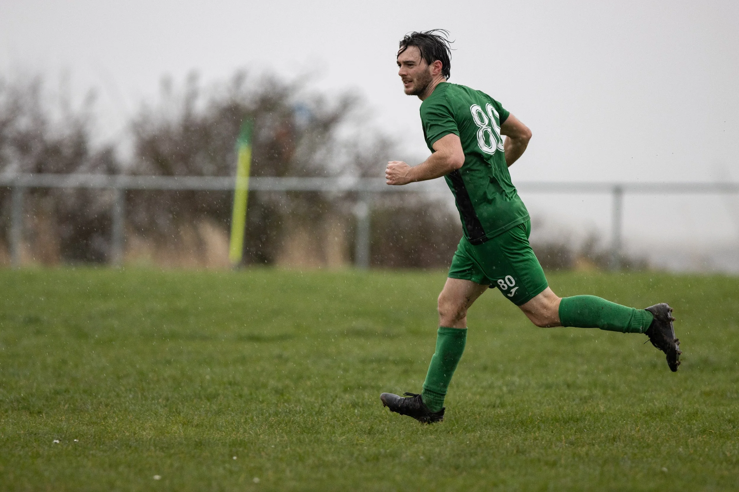 A soccer player wearing a green uniform running on a grassy field during a match in rainy weather.