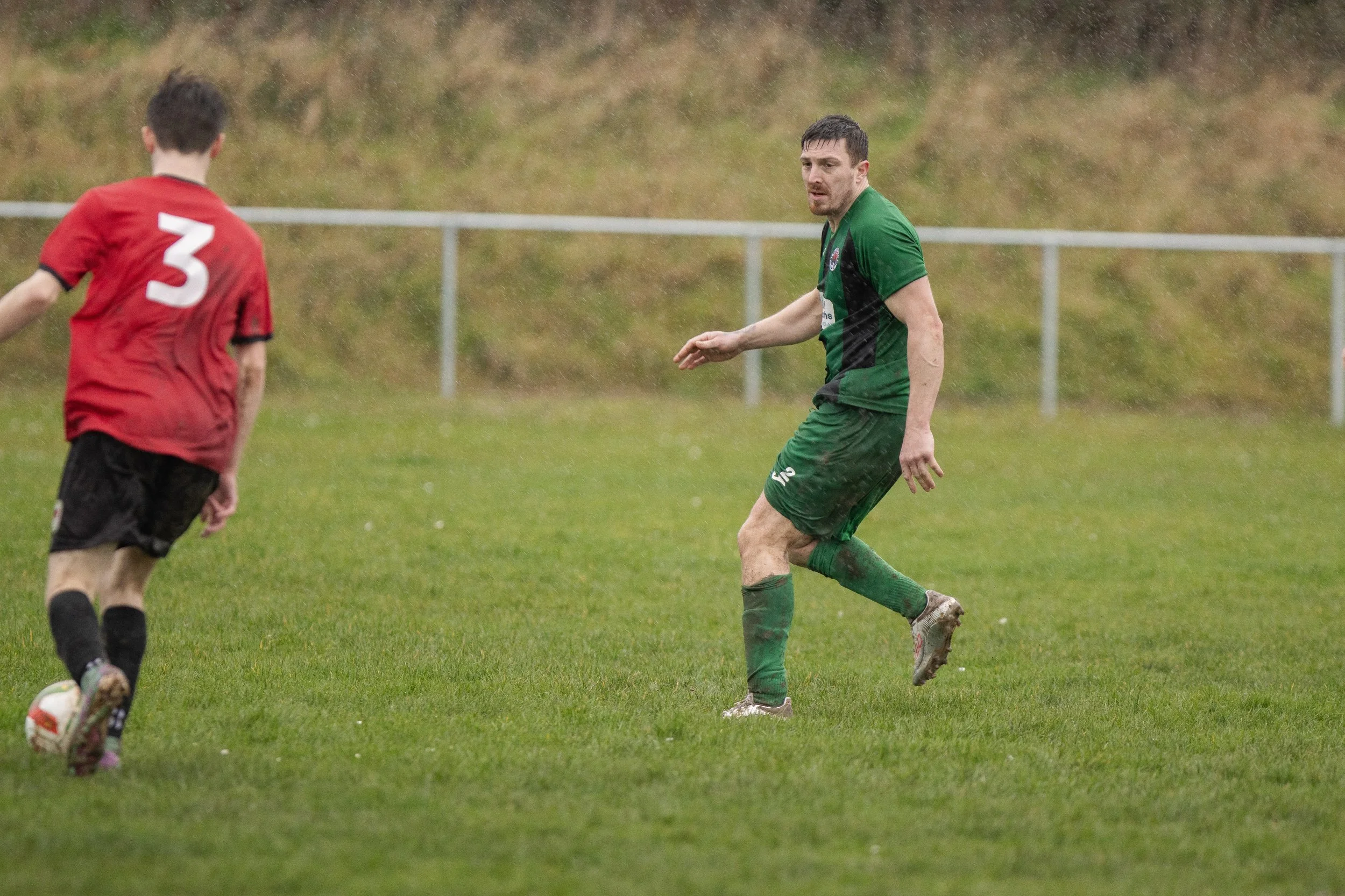 Two soccer players on a grassy field during a game in rainy weather, one wearing a red shirt with number 3 and black shorts, and the other in a green shirt and shorts with muddy socks and shoes.