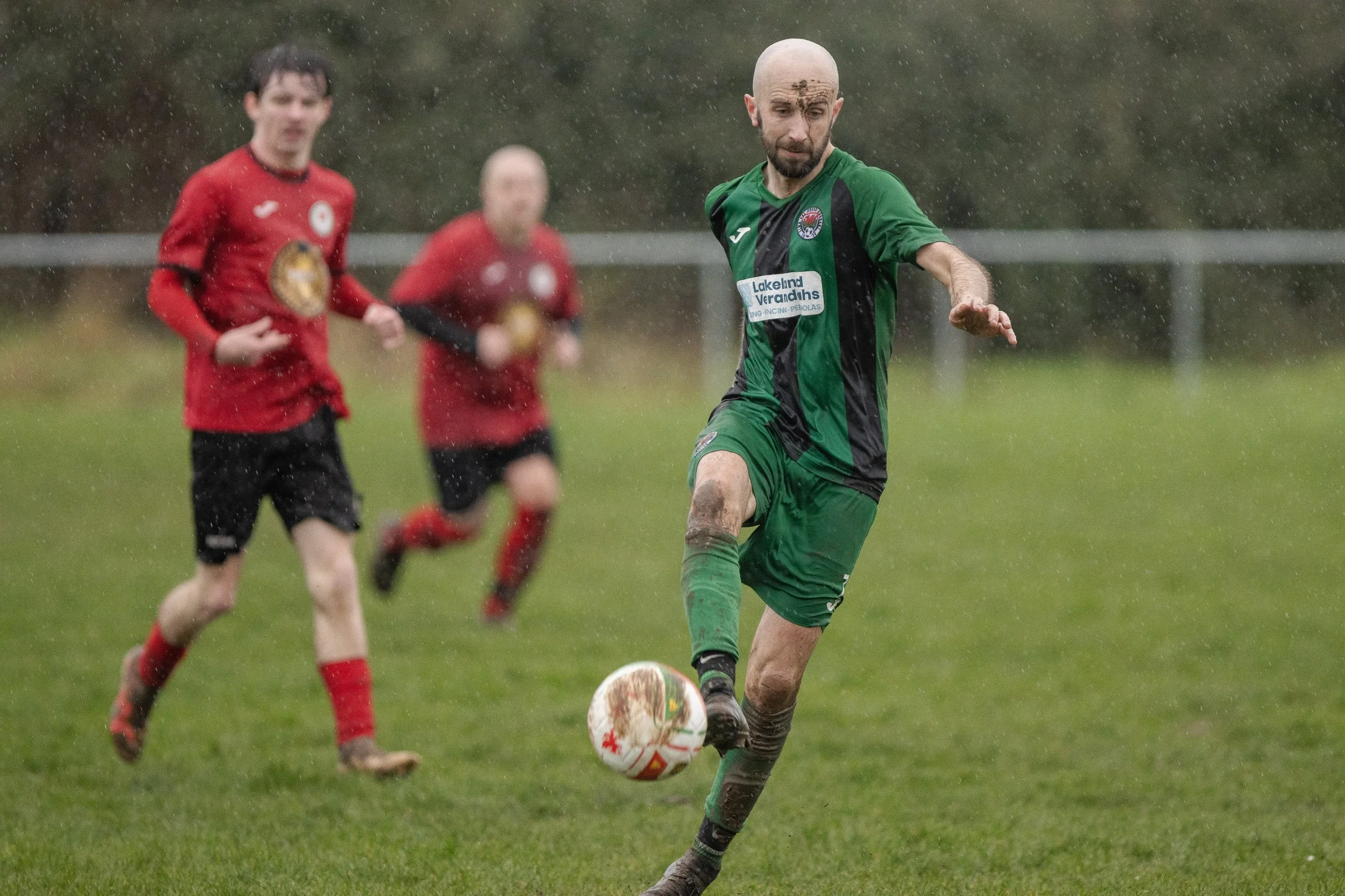 Soccer players playing in the rain on a grassy field, with one player in a green and black uniform kicking a ball while two players in red and black uniforms chase.