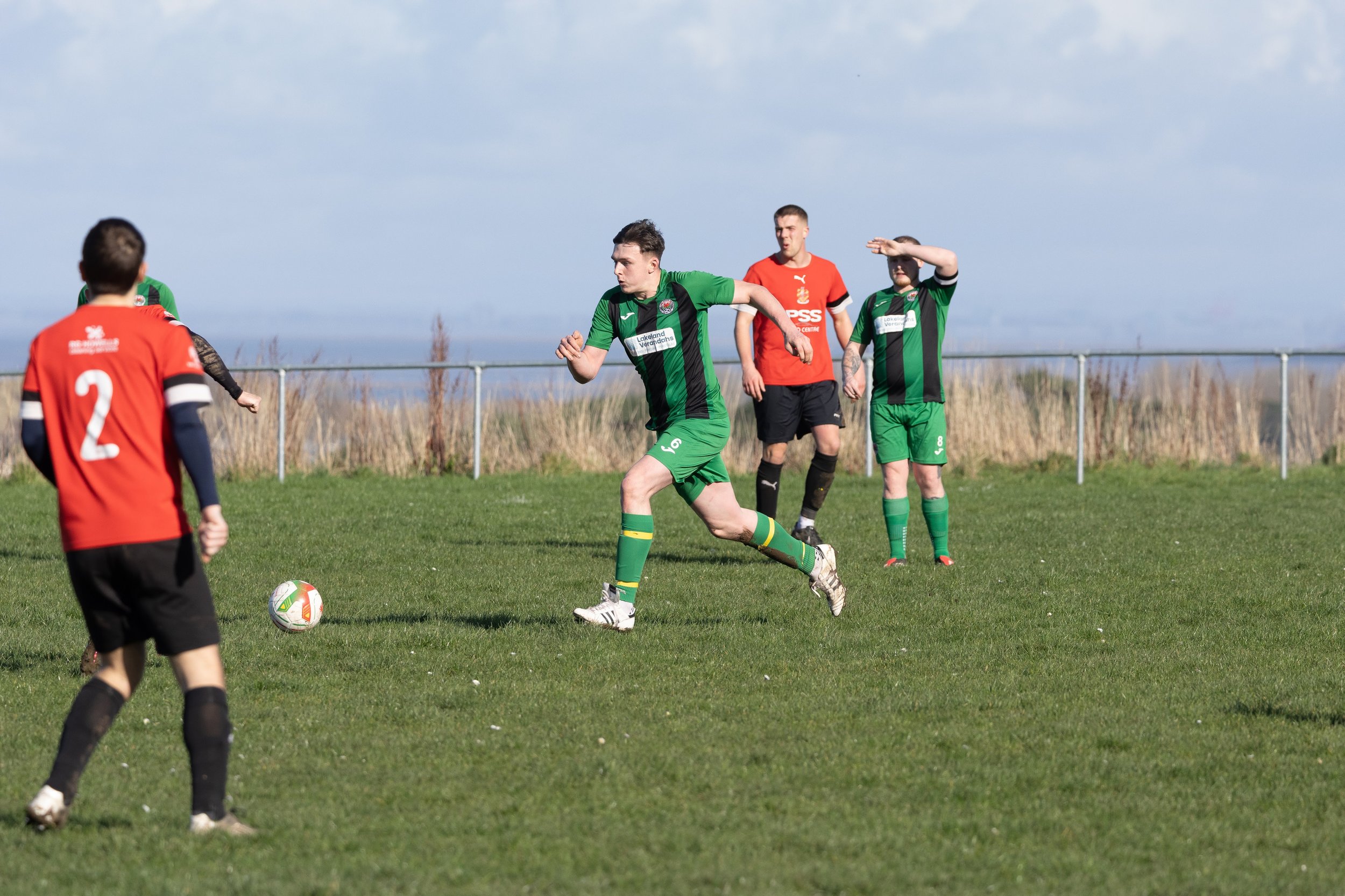 Young men playing soccer on a grass field, with some wearing green jerseys and one in a red jersey, under a cloudy sky.