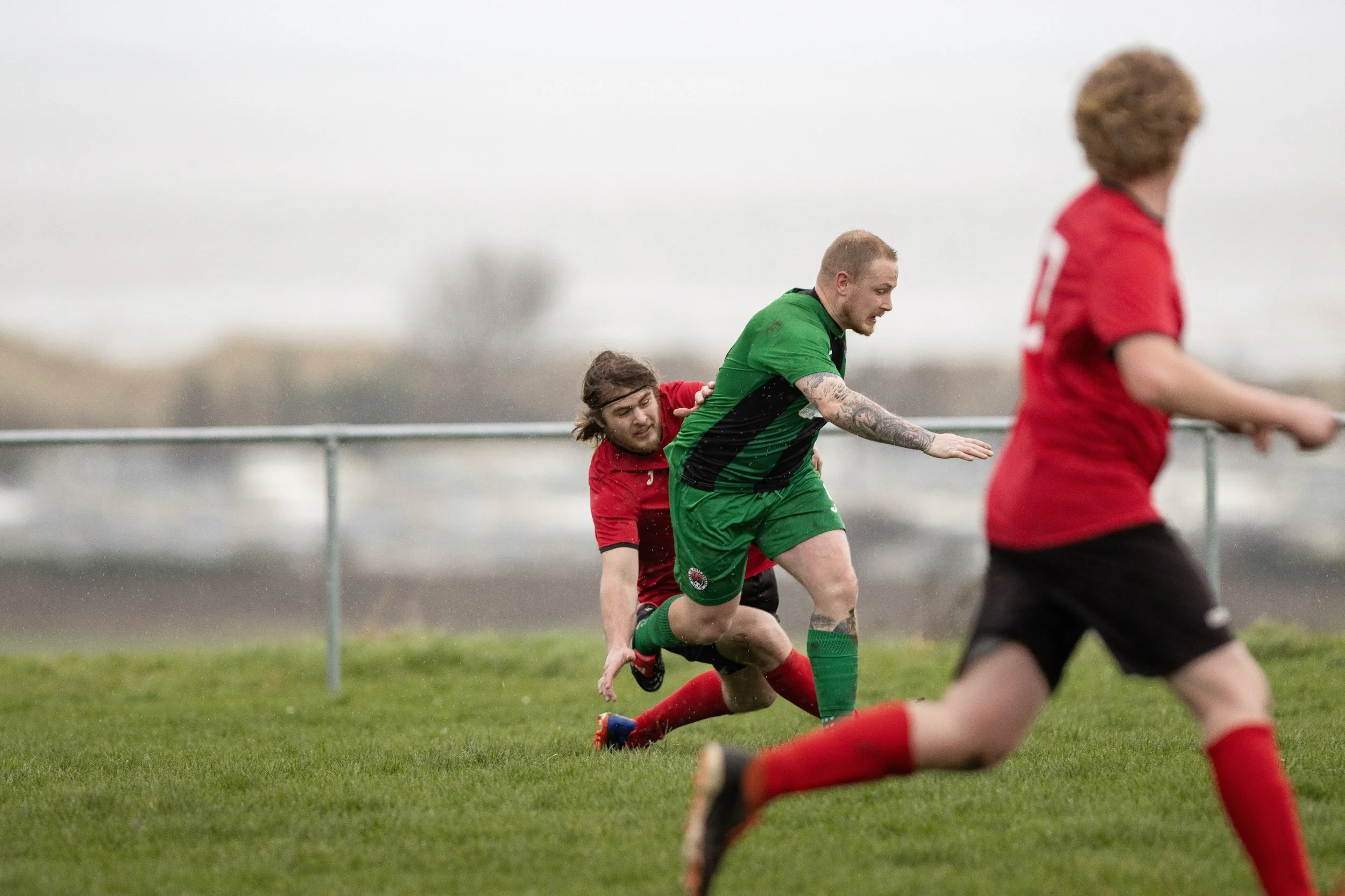 Two soccer players, one in red and one in green, competing for the ball on a grassy field in rainy weather. Another player in red is partly visible in the foreground.