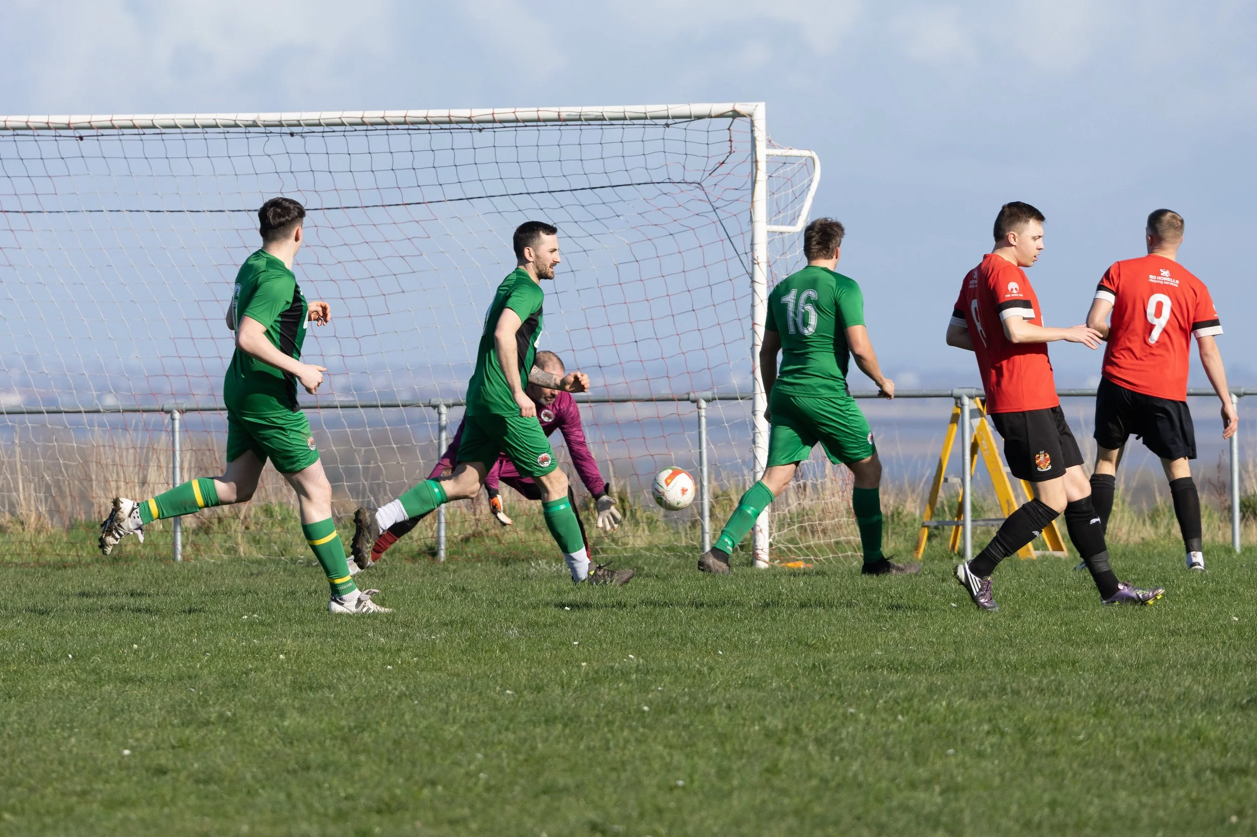 Soccer players in green and red jerseys near goal, with a goalkeeper in purple gloves, during a match on a grassy field.