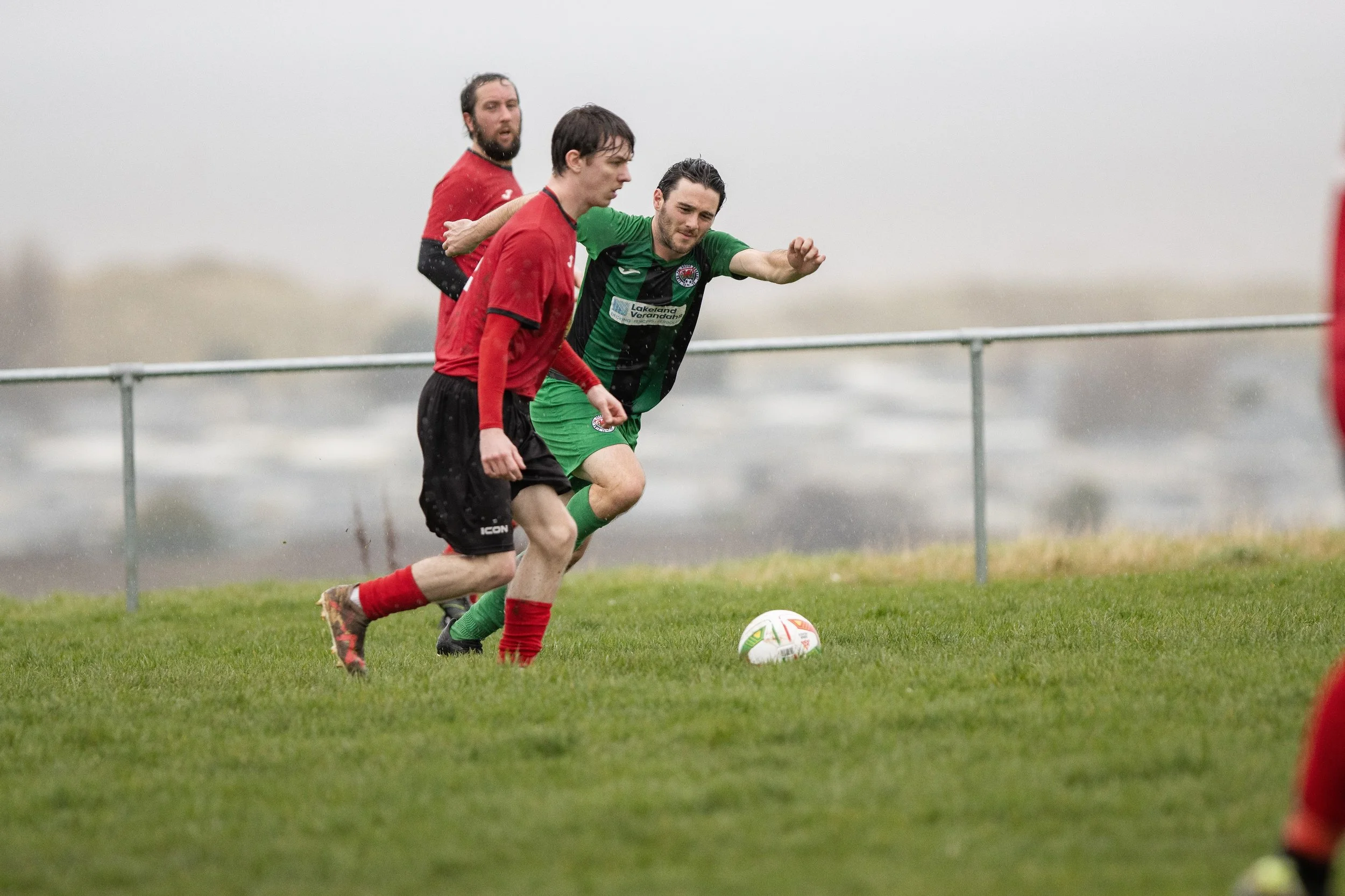 Two soccer players in red and green jerseys compete for possession of the ball on a grassy field during rainy weather.