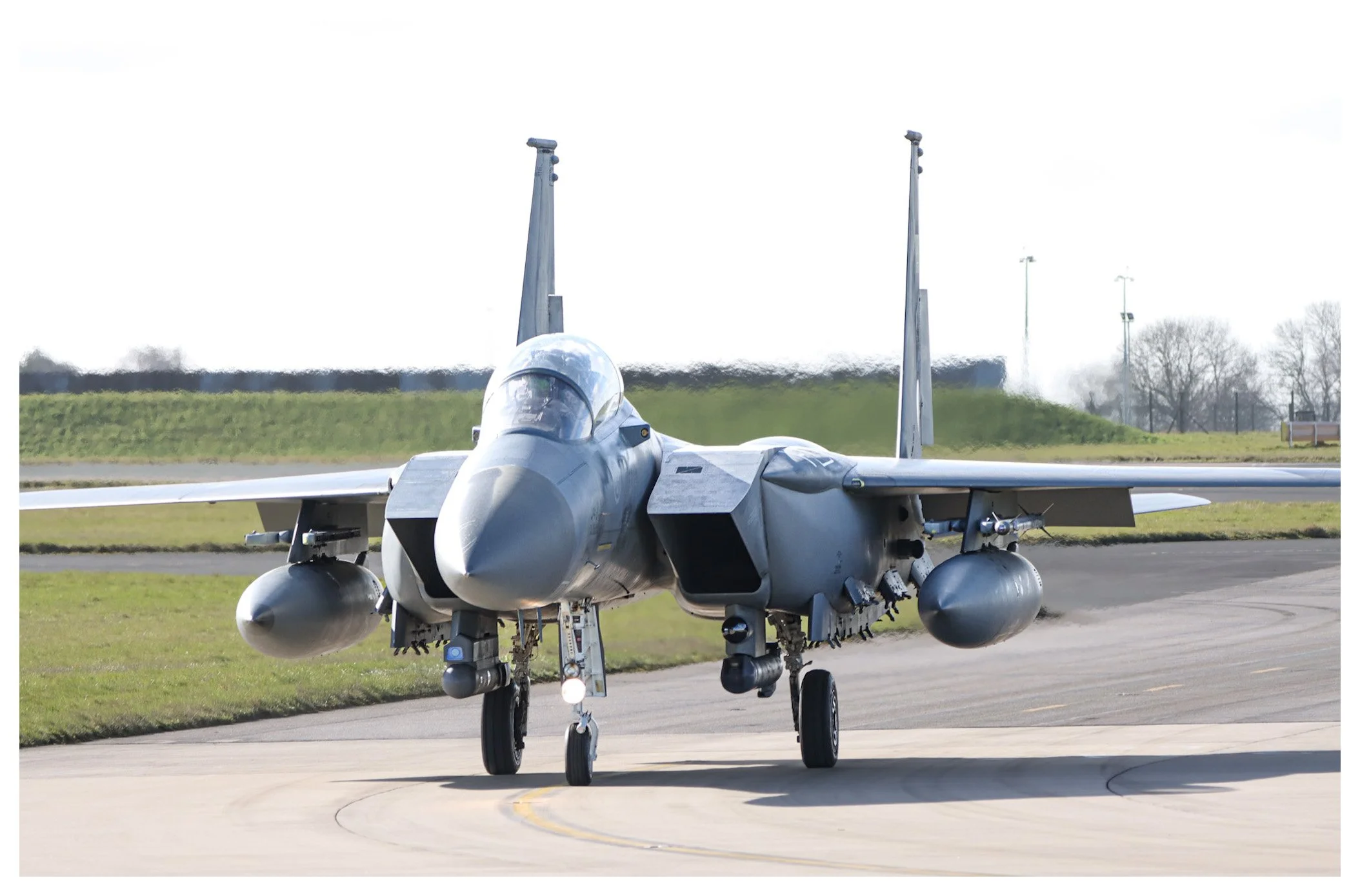 A military fighter jet parked on an airstrip with a grassy background and trees in the distance.