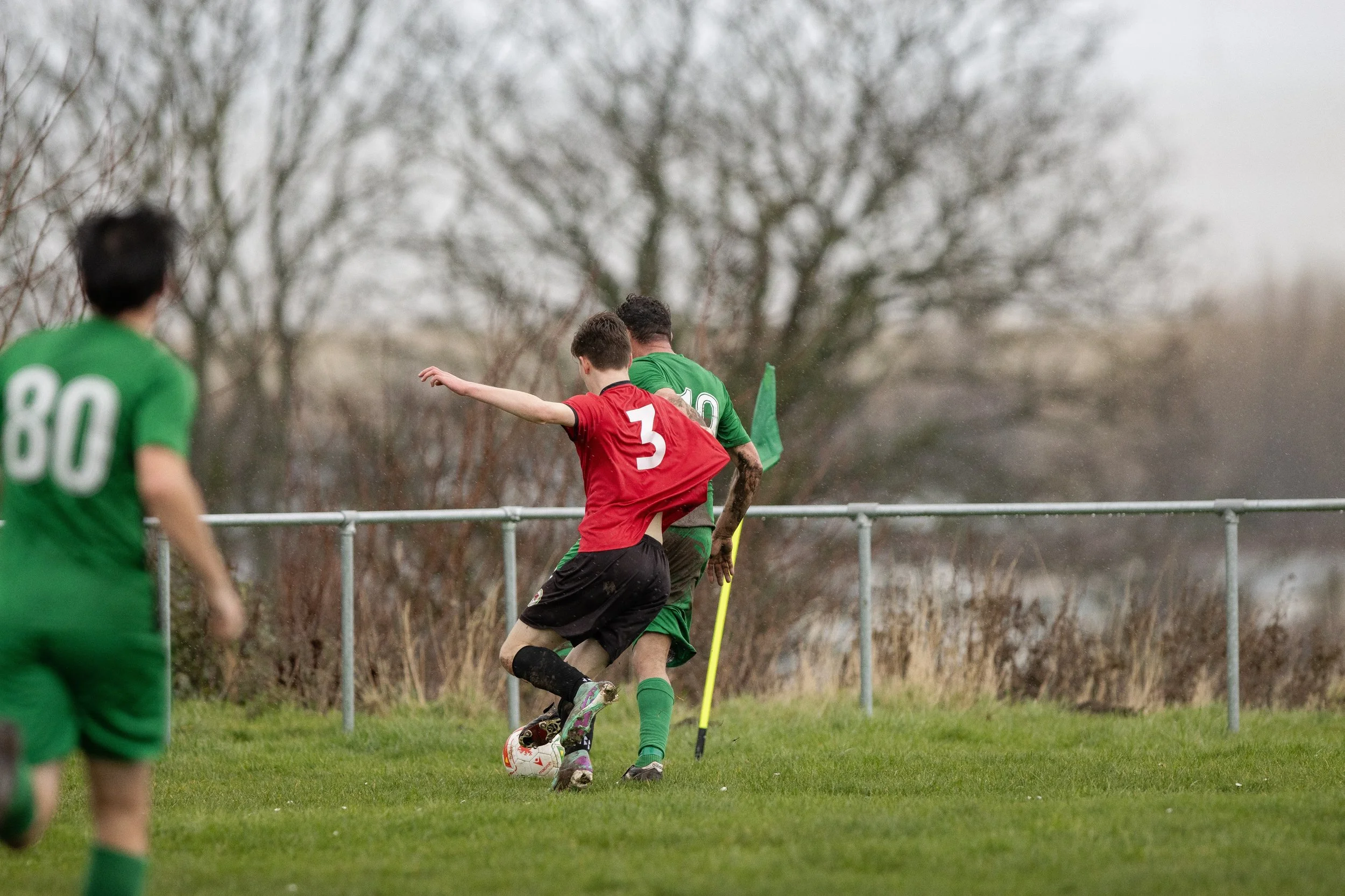 Young soccer players in green and red jerseys compete on a grassy field with trees in the background.