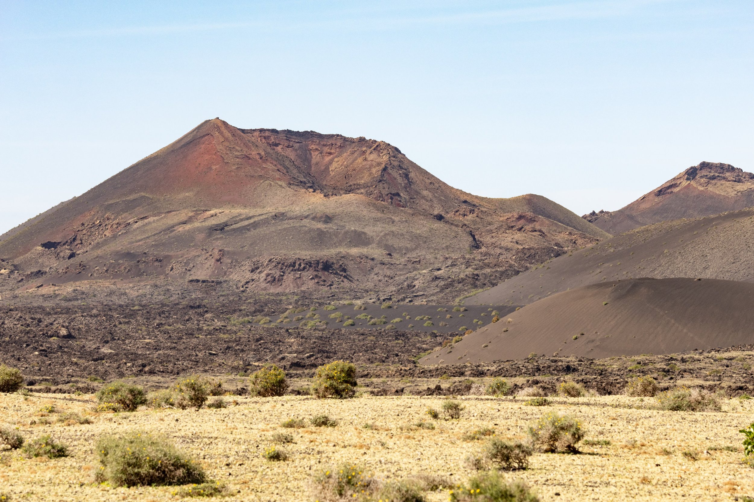 A desert landscape with dark volcanic mountains, sparse vegetation, and a clear sky.