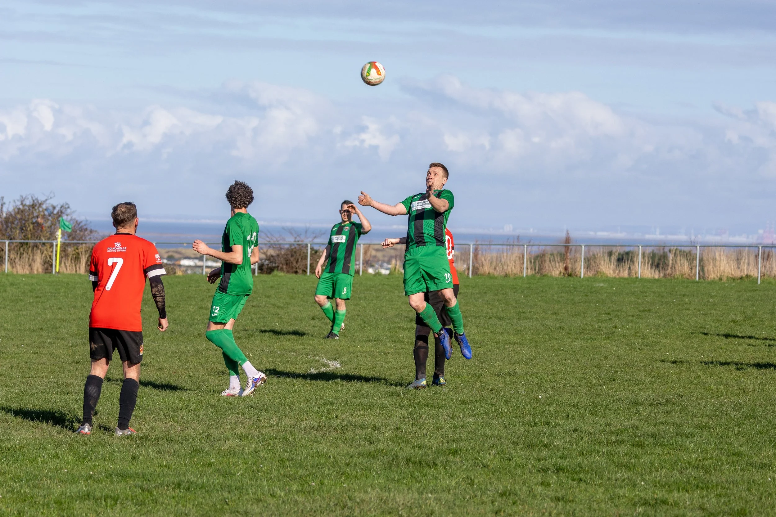 Soccer players on the field, with two players jumping to head the ball during a game on a grassy field with a fence and cloudy sky in the background.
