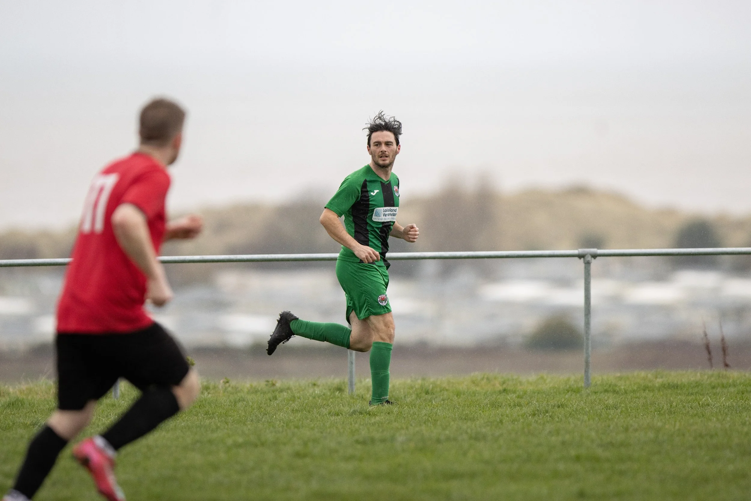 Two soccer players, one in red and one in green, running on a grassy field with a gray sky and blurry landscape in the background.