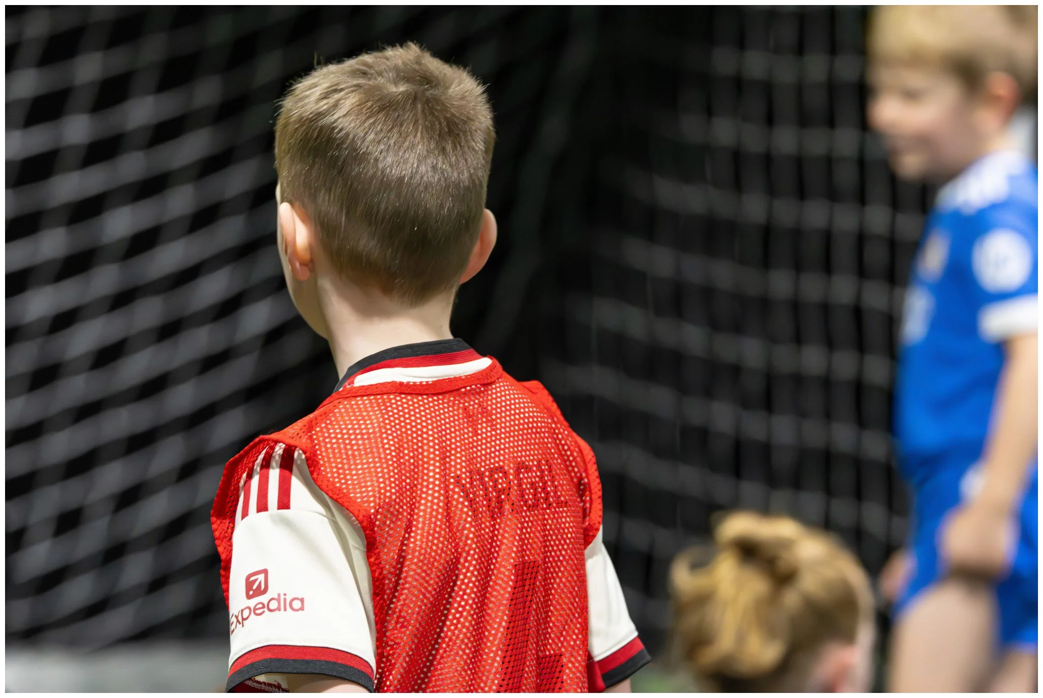 Young boy with short light brown hair seen from behind, wearing a red and white sports jersey with a black collar, standing in front of a black netting, possibly on a sports field.