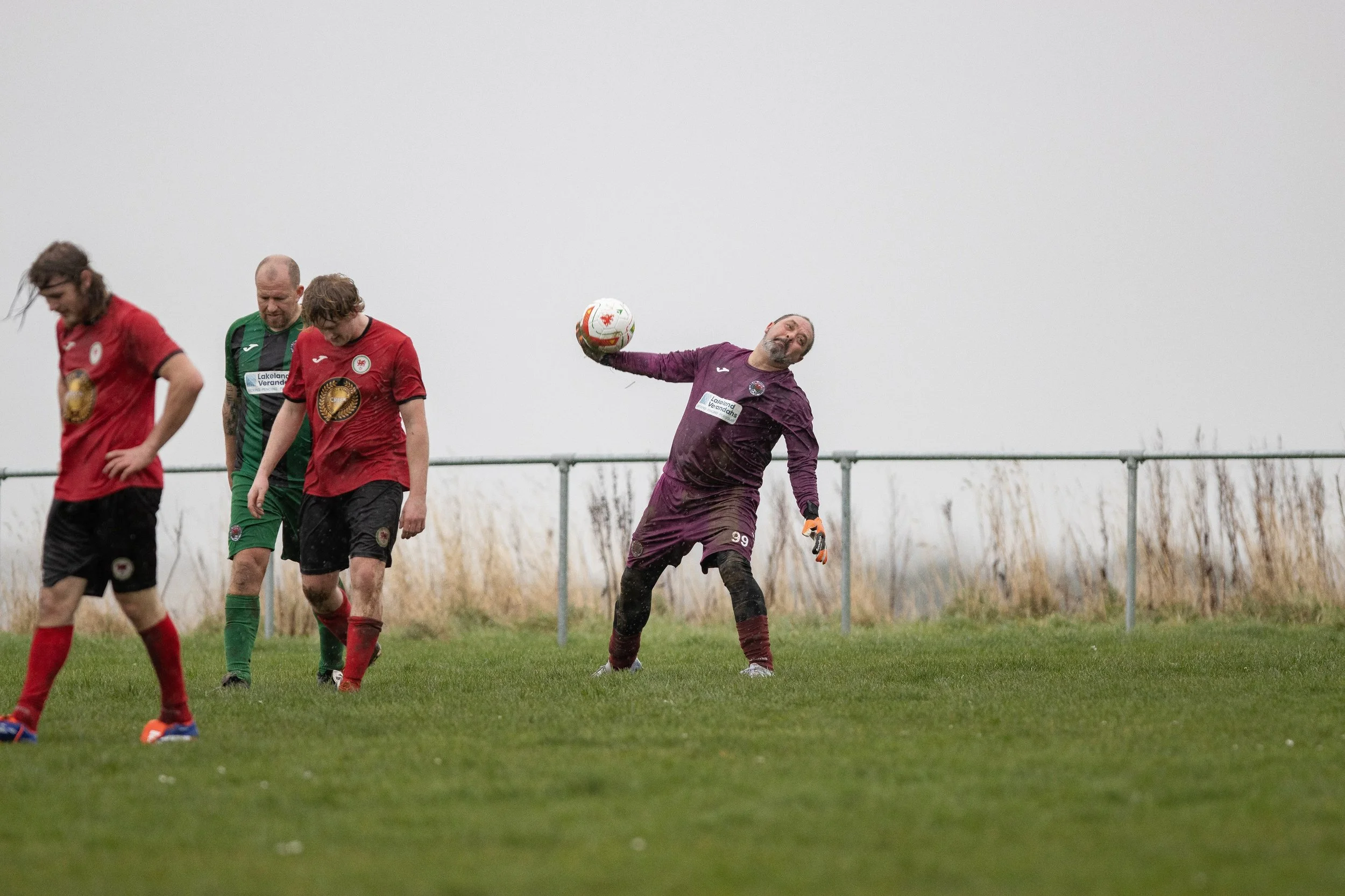 A soccer game on a rainy day with four players on the field, including a goalkeeper in purple making a save, while three outfield players in red and green look on, all with wet and muddy appearances.