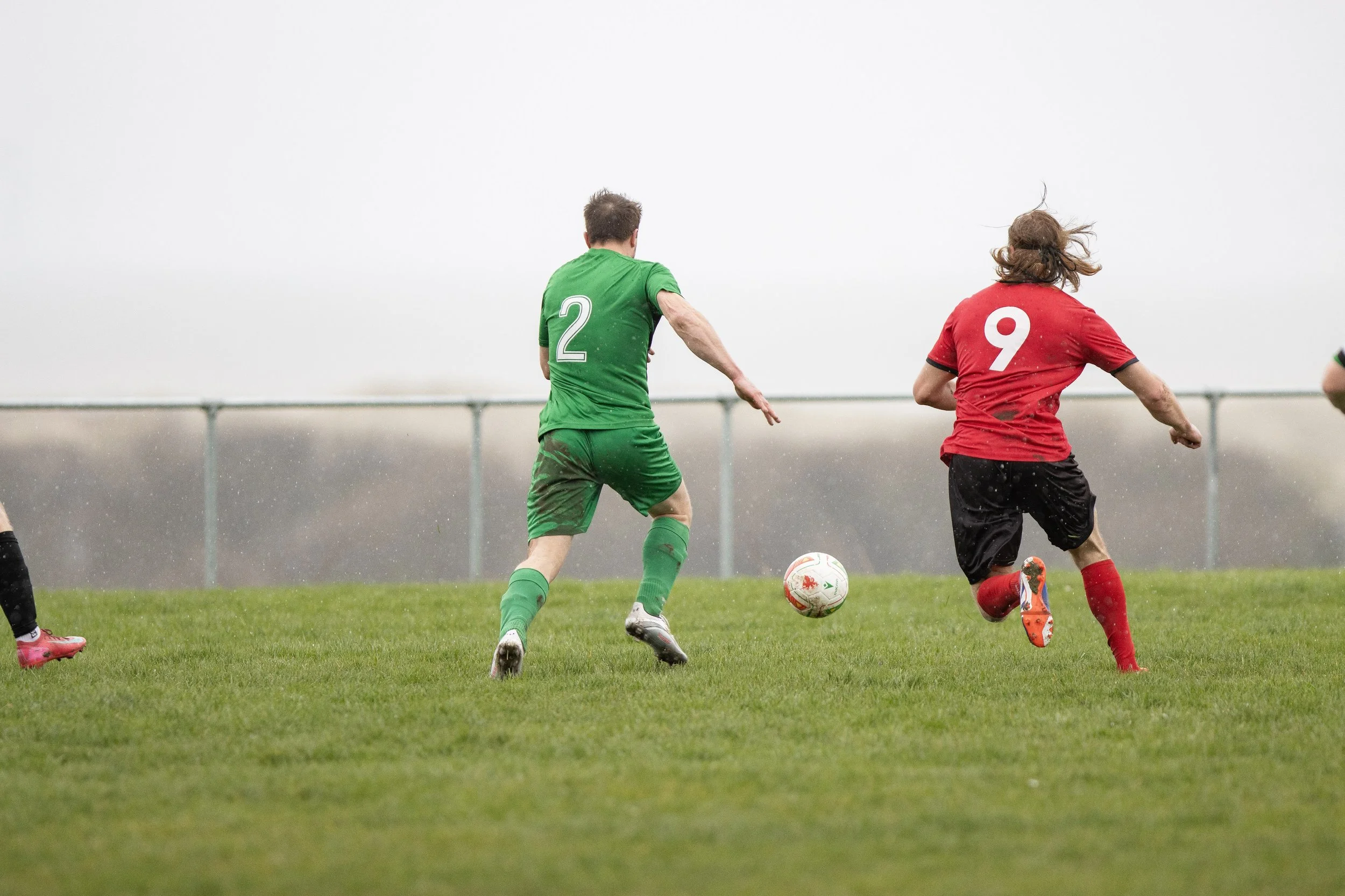 Two soccer players, one in a green uniform with number 2 and the other in a red uniform with number 9, chasing a soccer ball on a grass field in overcast weather.
