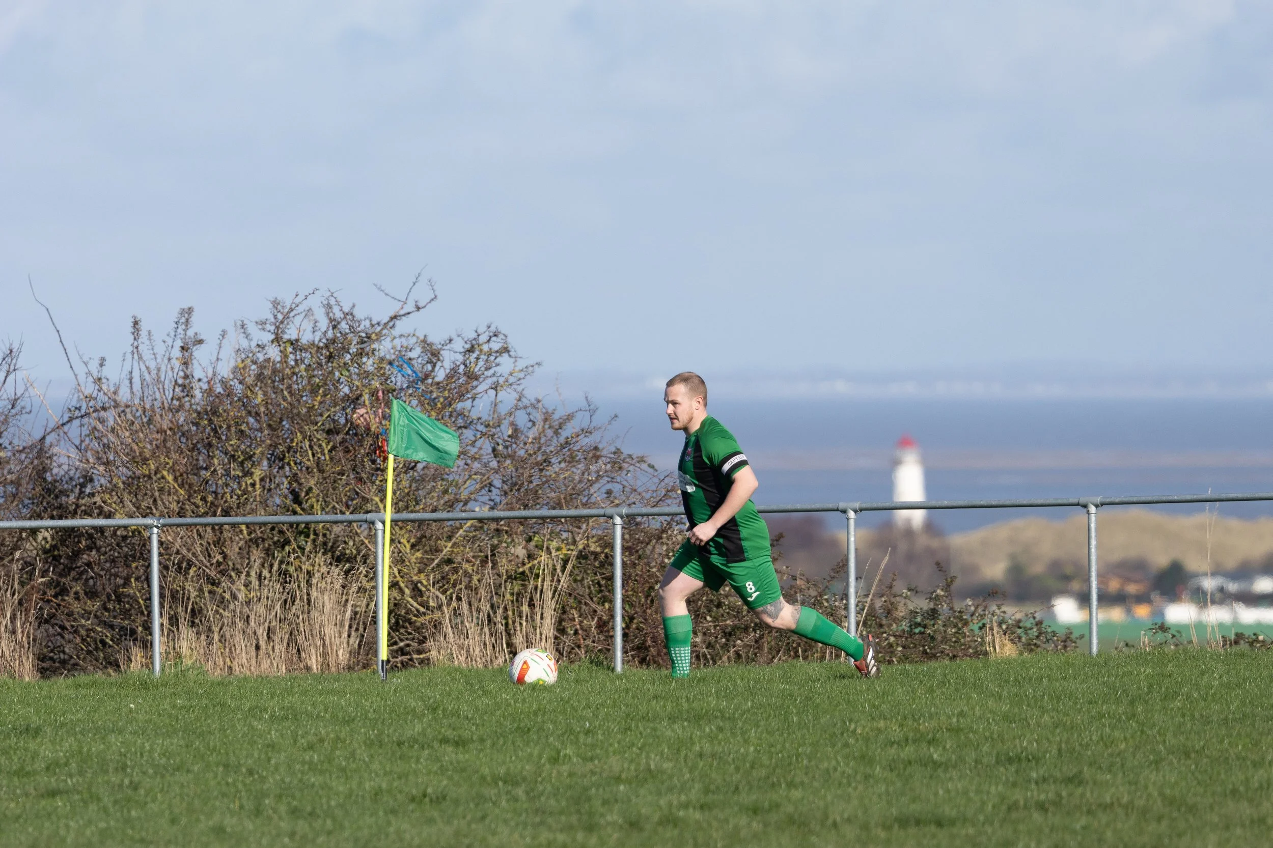 A soccer player in a green uniform running on the field towards a soccer ball with a lighthouse visible in the background.