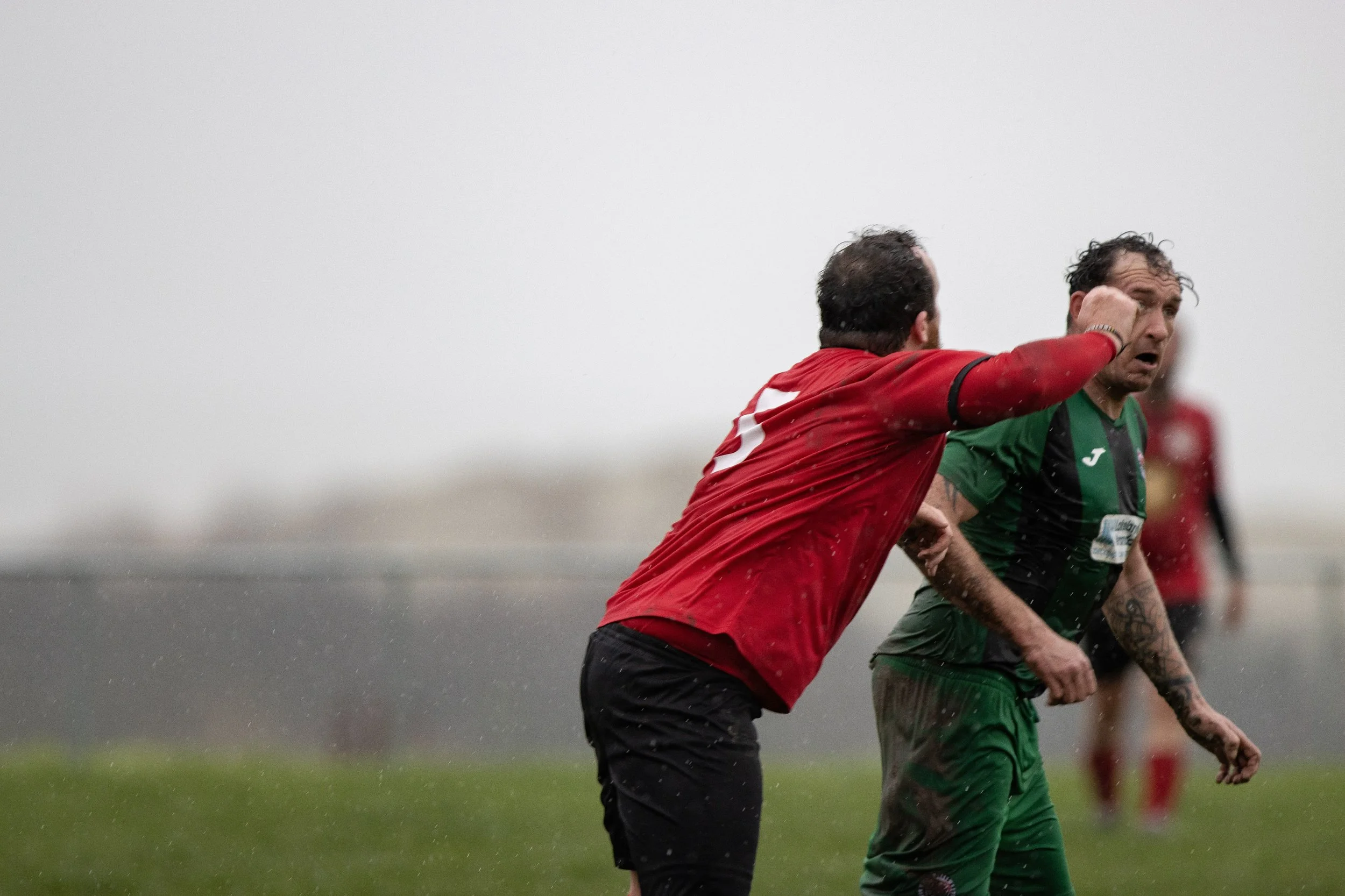 A soccer referee in a red shirt appears to be hitting a soccer player in a green uniform in the face during a match in rainy weather.