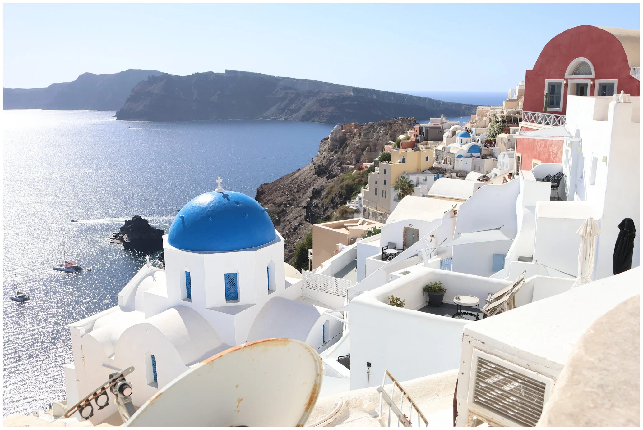 Scenic view of Santorini, Greece, showing white buildings with blue domes on a hillside overlooking the sea with boats, cliffs, and the ocean in the background.
