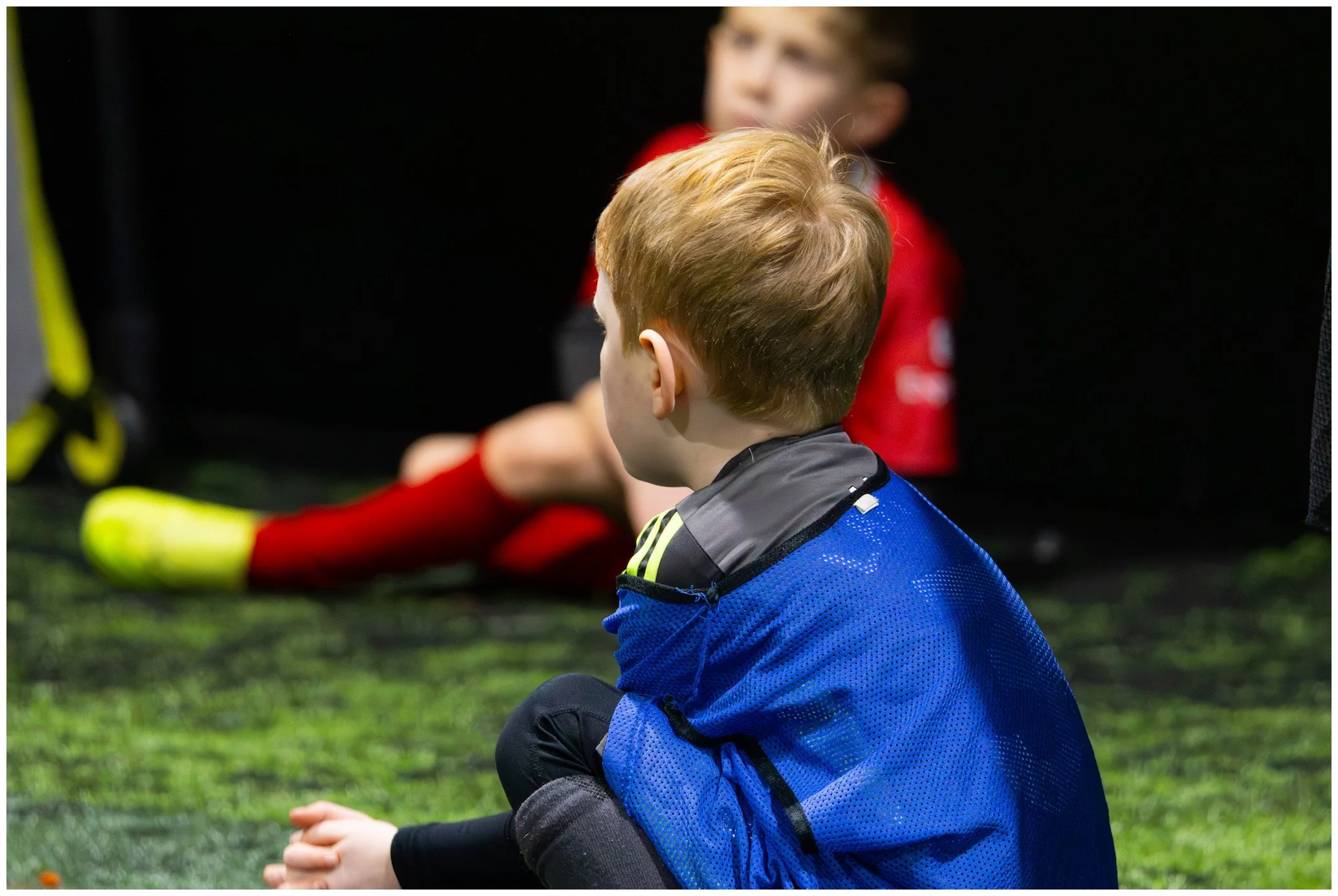 Young boy sitting on green artificial turf, observing a soccer game or practice, with a blurred soccer player in red uniform in the background.