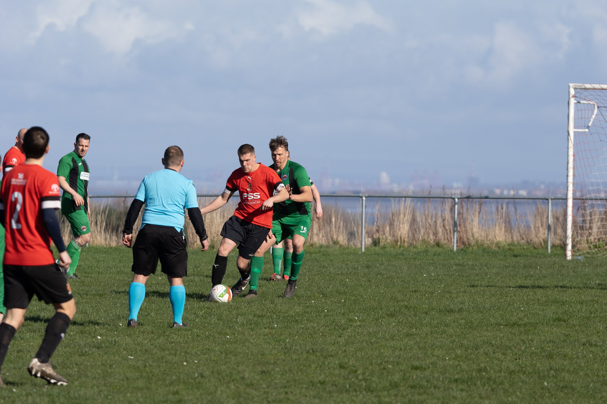 Soccer players in red and green uniforms contest a ball on a grassy field near a goal, with a referee in a blue shirt observing; cloudy sky and open landscape in the background.