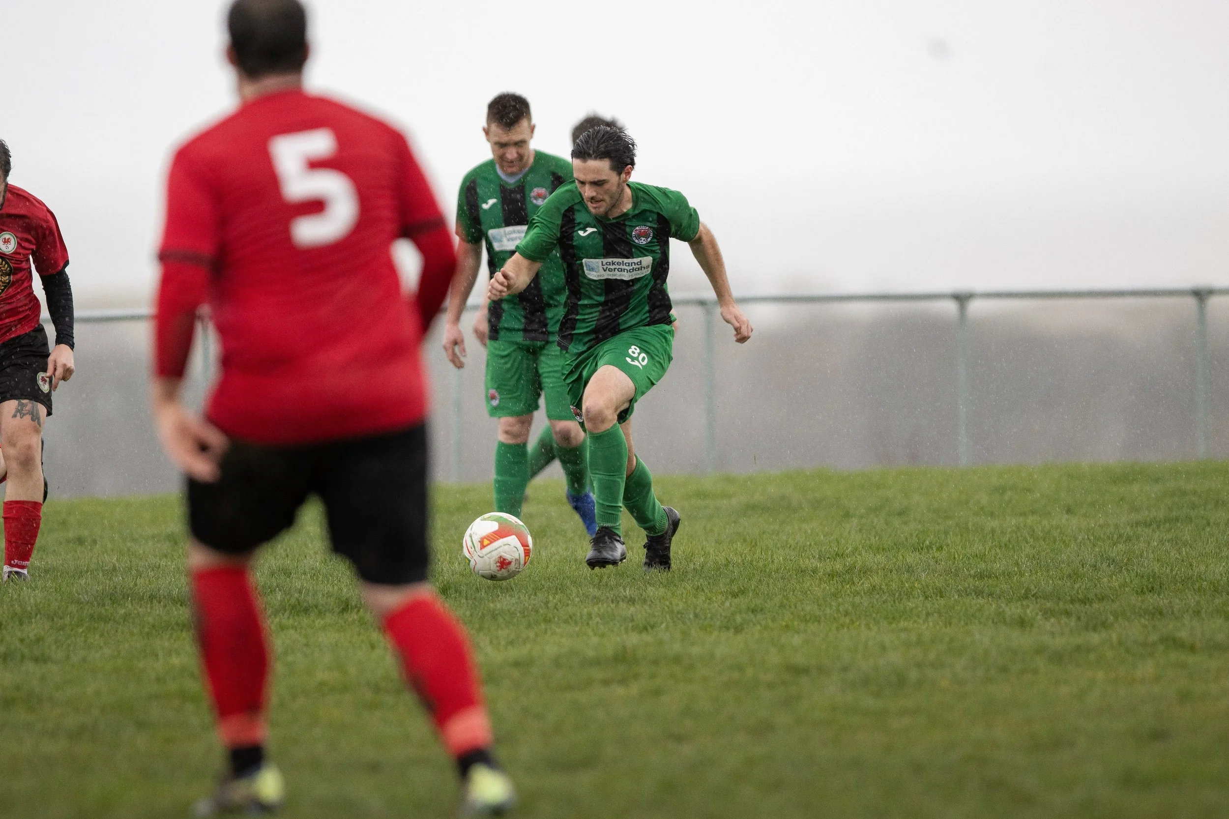 Soccer players in green and red uniforms playing on a grassy field in rainy weather