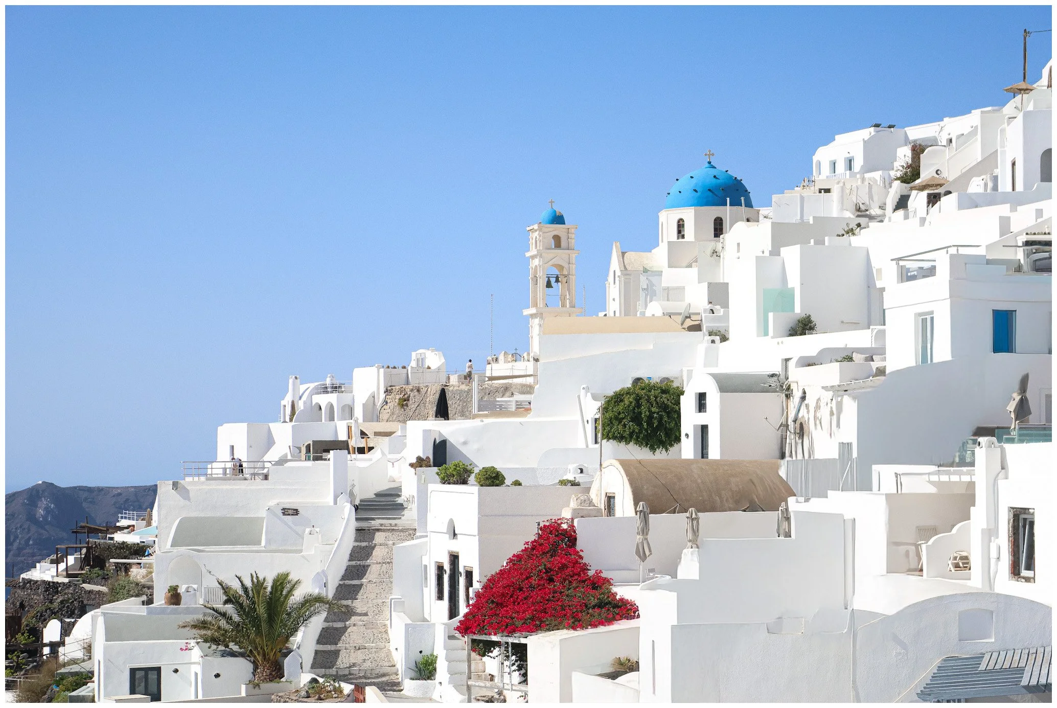 White buildings with blue domes on a hillside, typical of Santorini, Greece, under a clear blue sky.