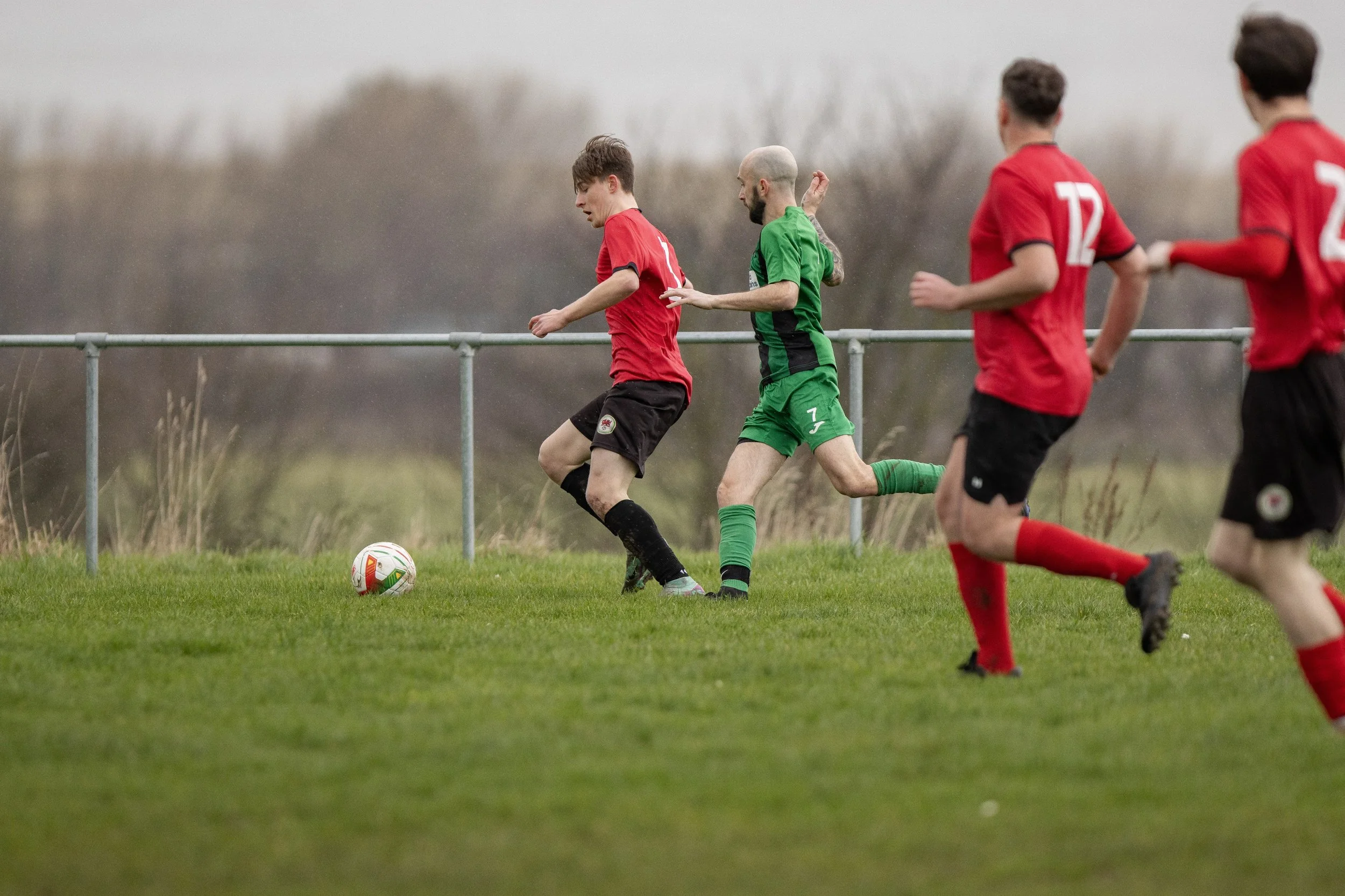 Soccer players on the field, with two players from opposing teams chasing the ball near a metal fence, during daytime with overcast sky.