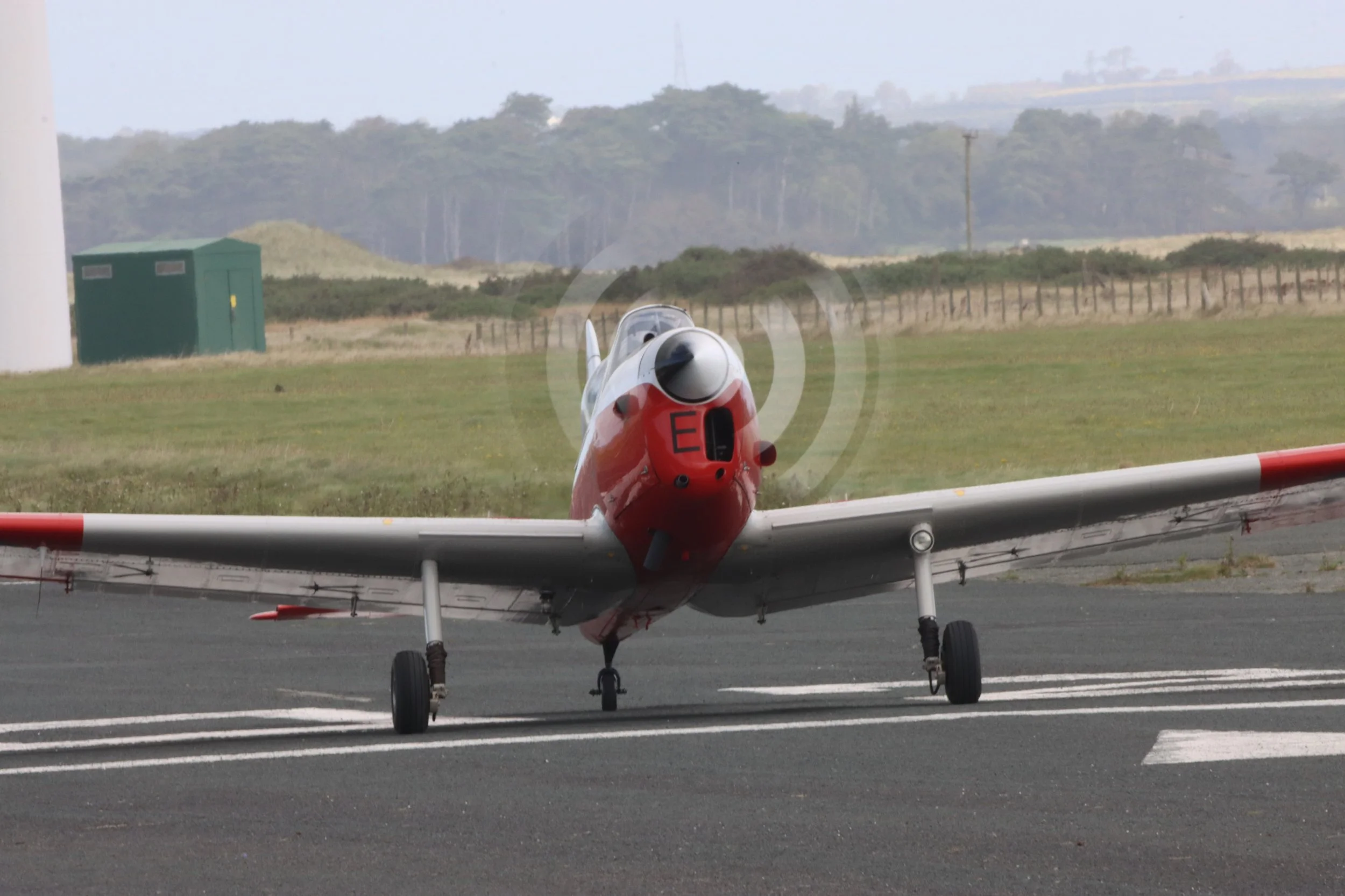 A vintage red and white propeller airplane on the runway, facing the camera with mountains and green fields in the background.