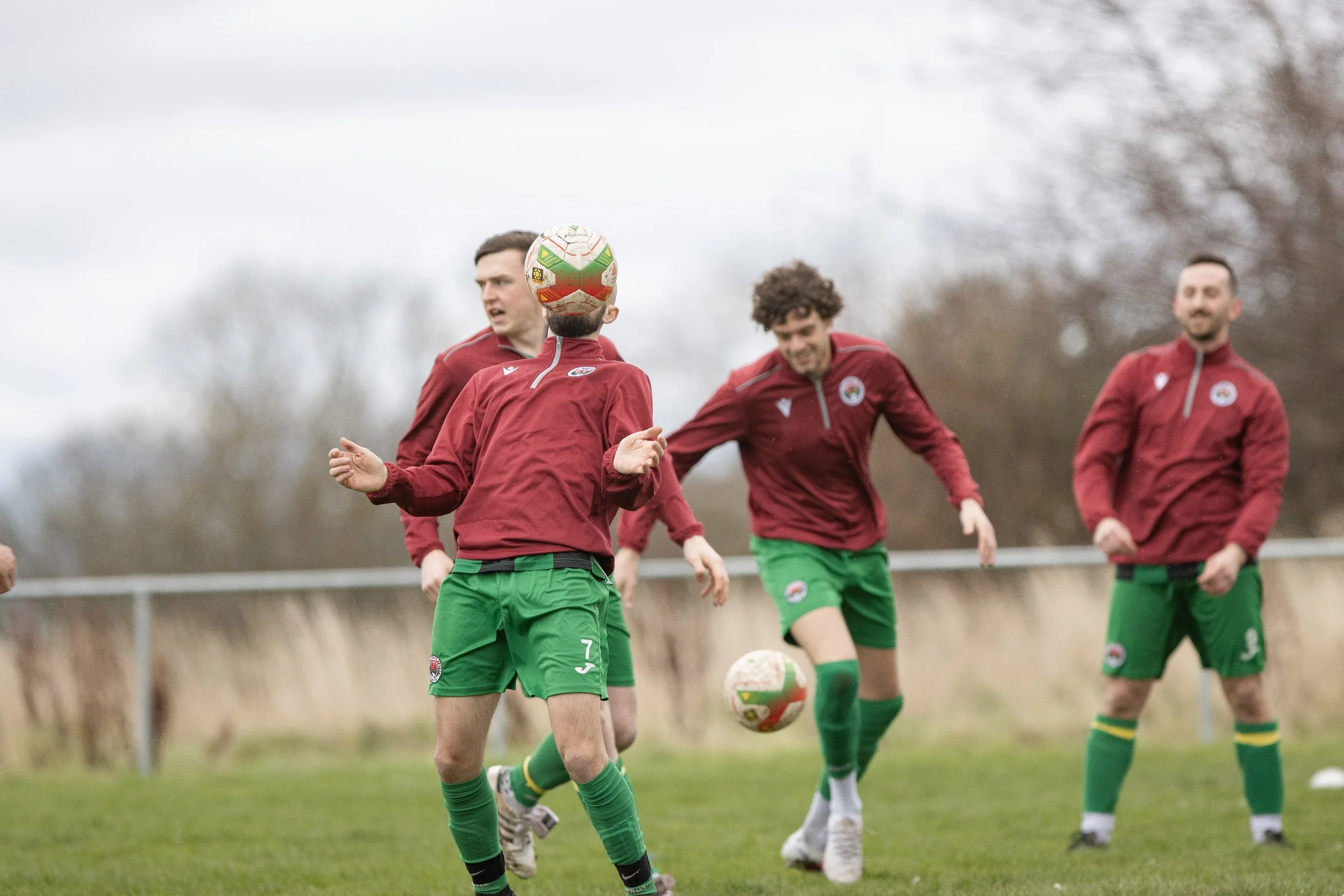 Soccer players practicing on a field, wearing maroon jackets and green shorts, with one ball in mid-air.
