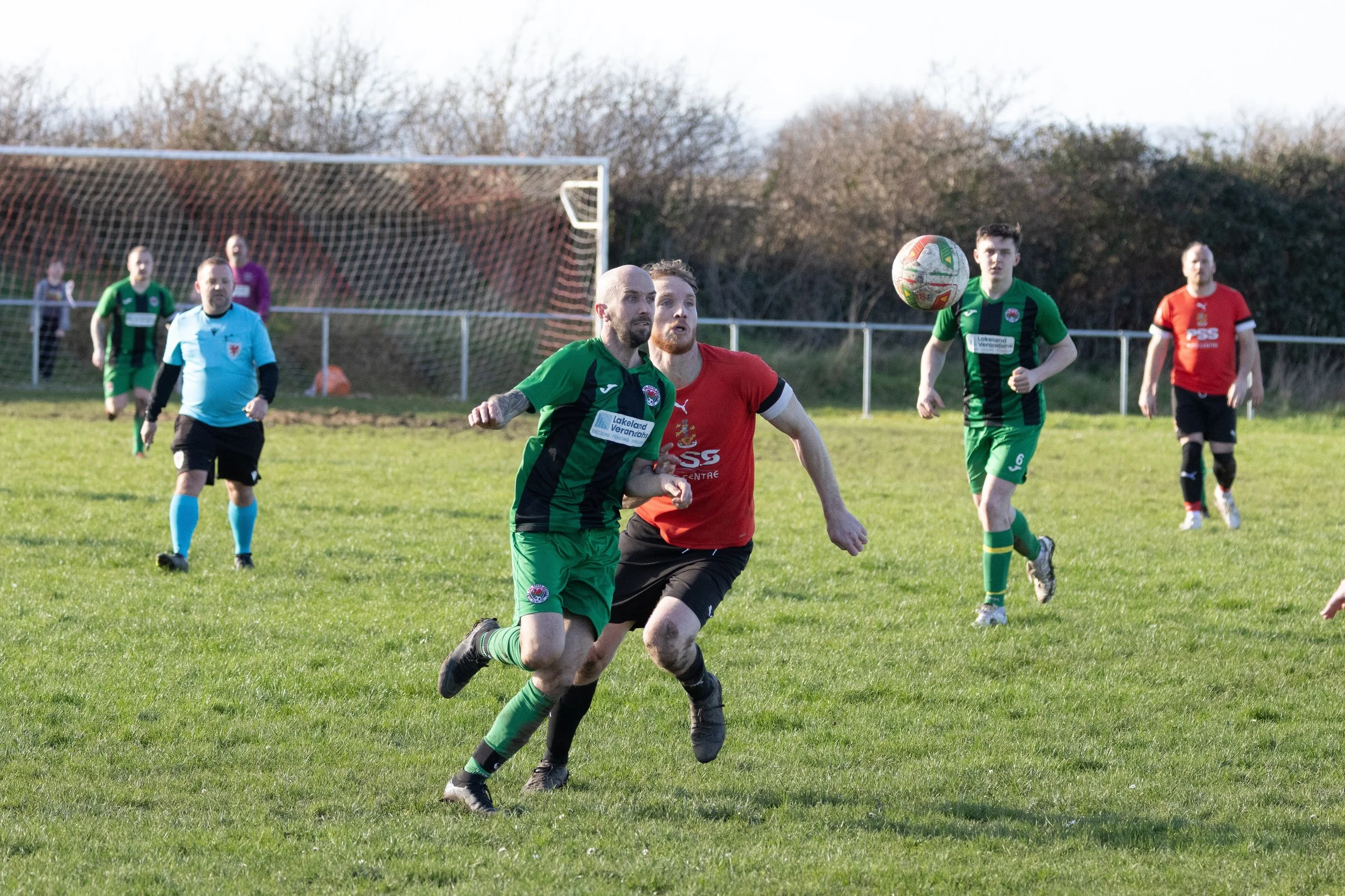 Soccer match with players running after the ball on a grassy field, goalpost in background, some players in green jerseys, one in red, and a referee in light blue with black shorts.